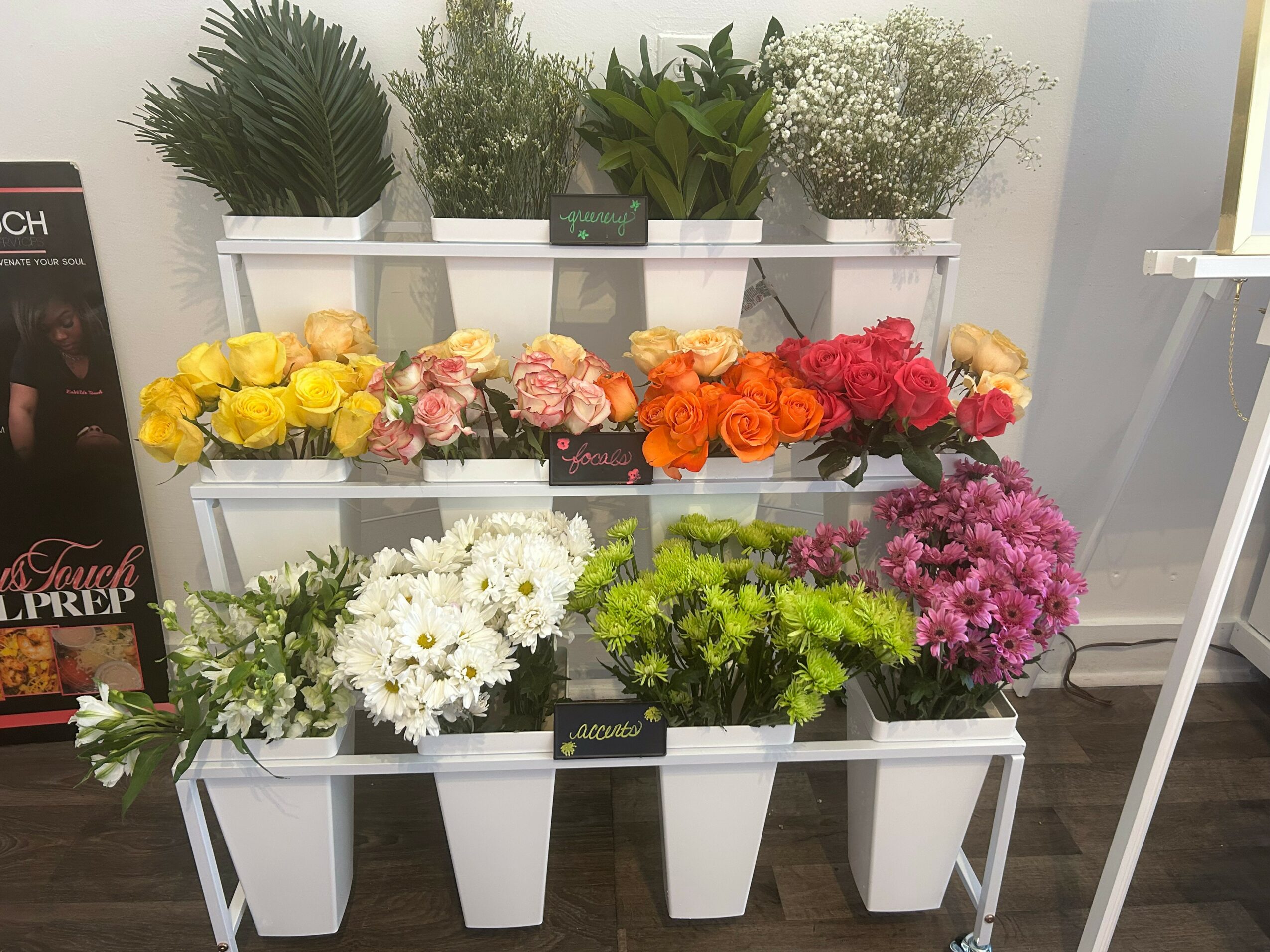 White plant stand with three shelves holding various potted plants and colorful flowers, set against a light-colored wall.