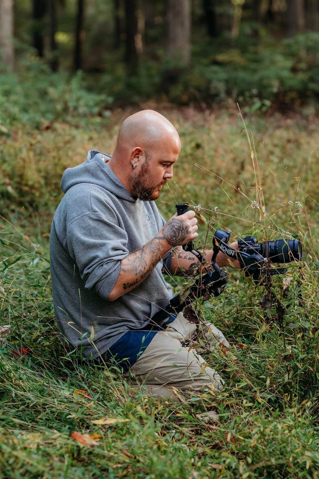 Man with a beard in a gray hoodie sitting in a forest, holding a camera and a smartphone, surrounded by greenery.