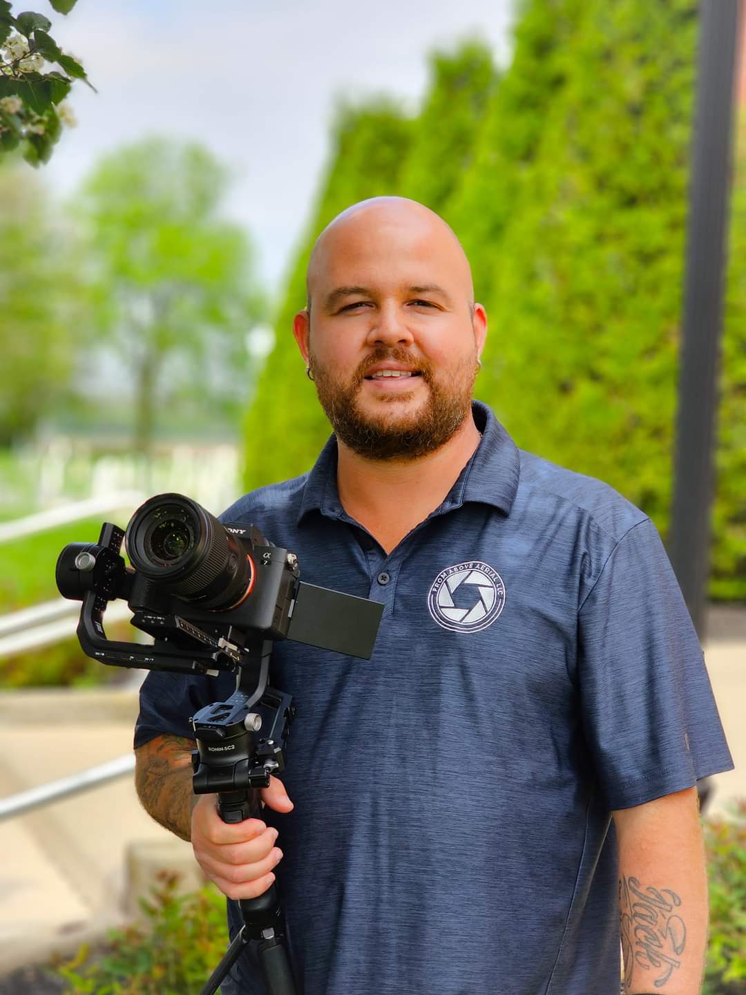 Man holding a camera on a tripod outdoors with green trees in the background.