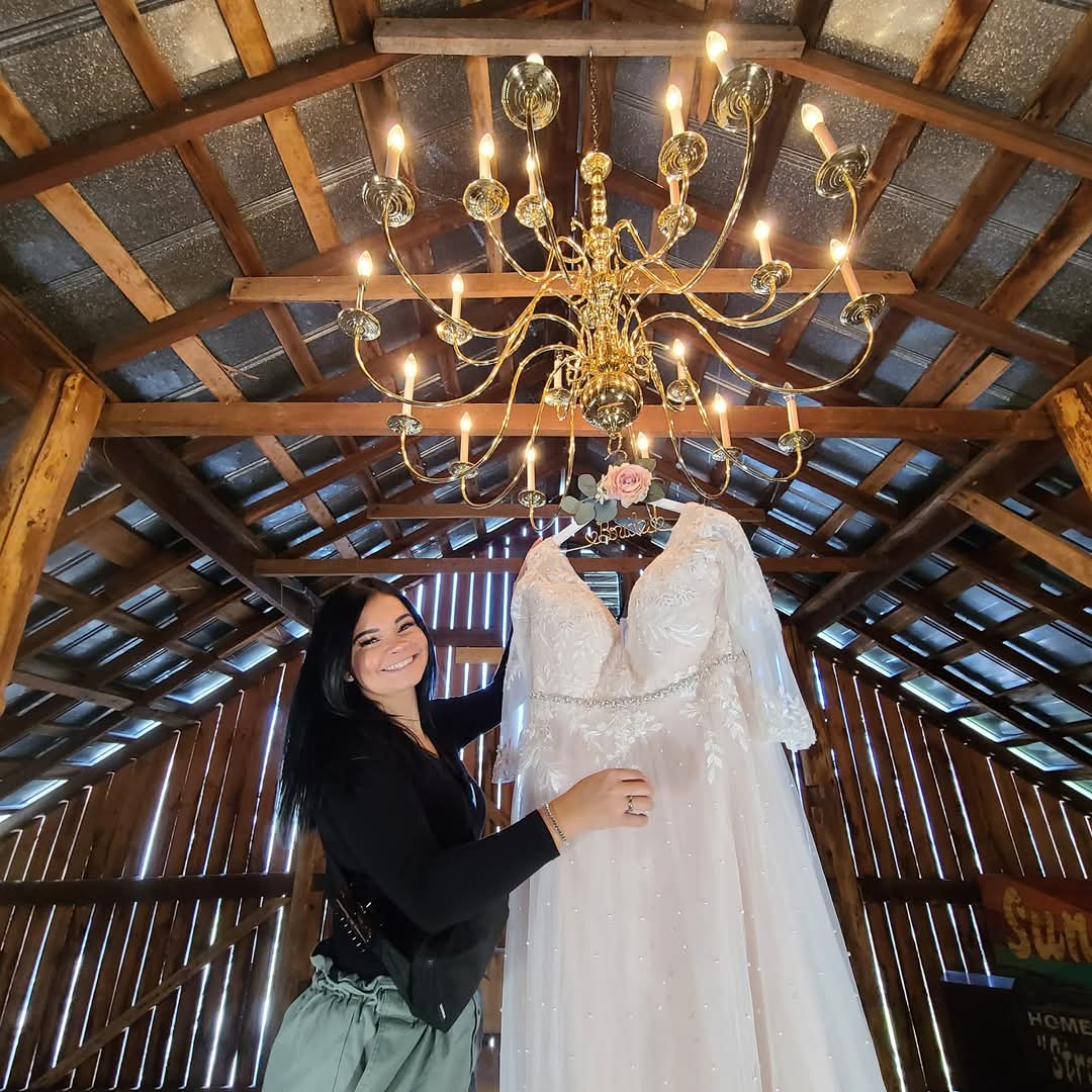 Woman in black top holding a white wedding dress hanging from a chandelier inside a wooden barn.