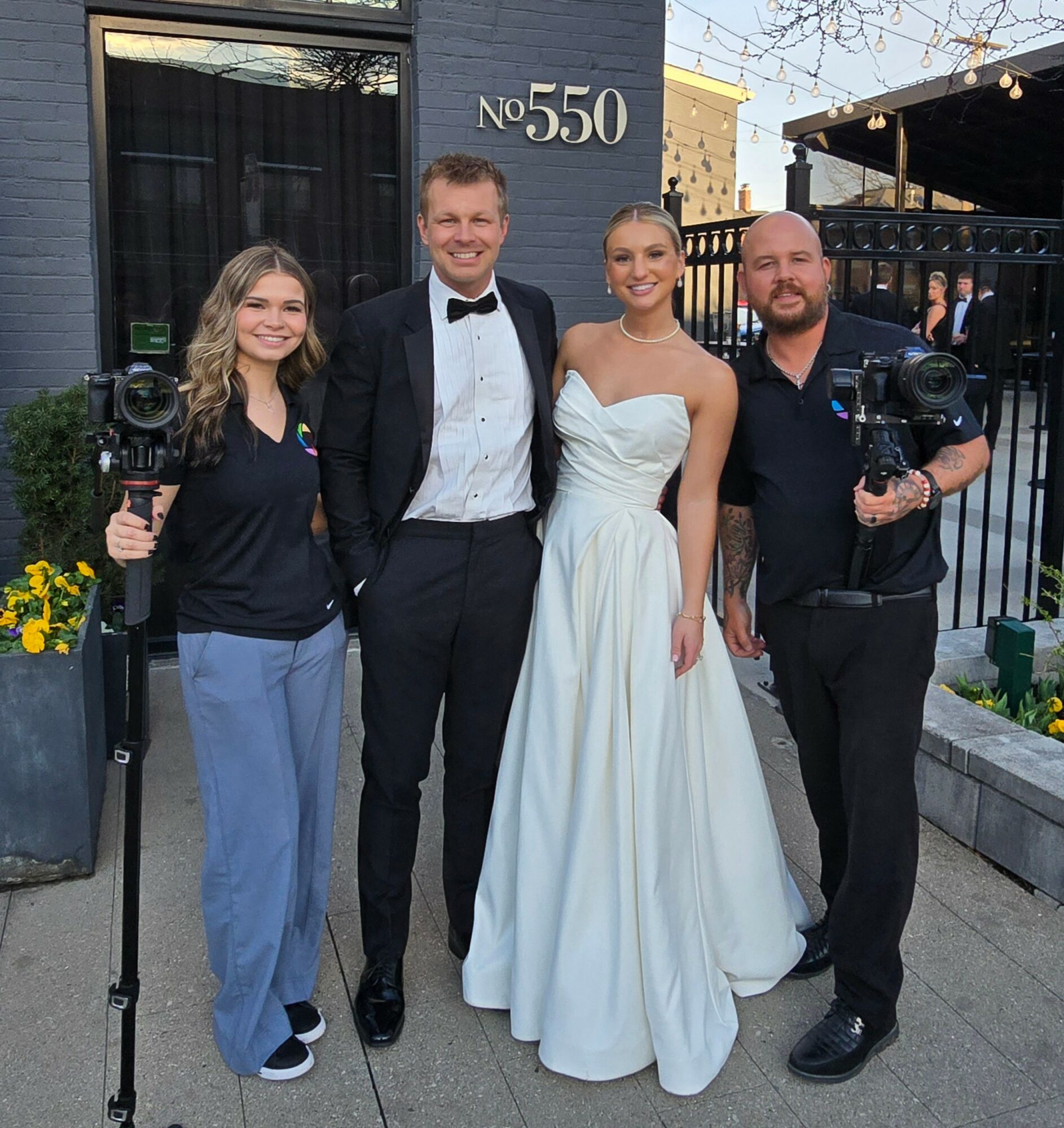 Four people standing outside a building, two holding cameras, one in a tuxedo, one in a white dress, smiling.