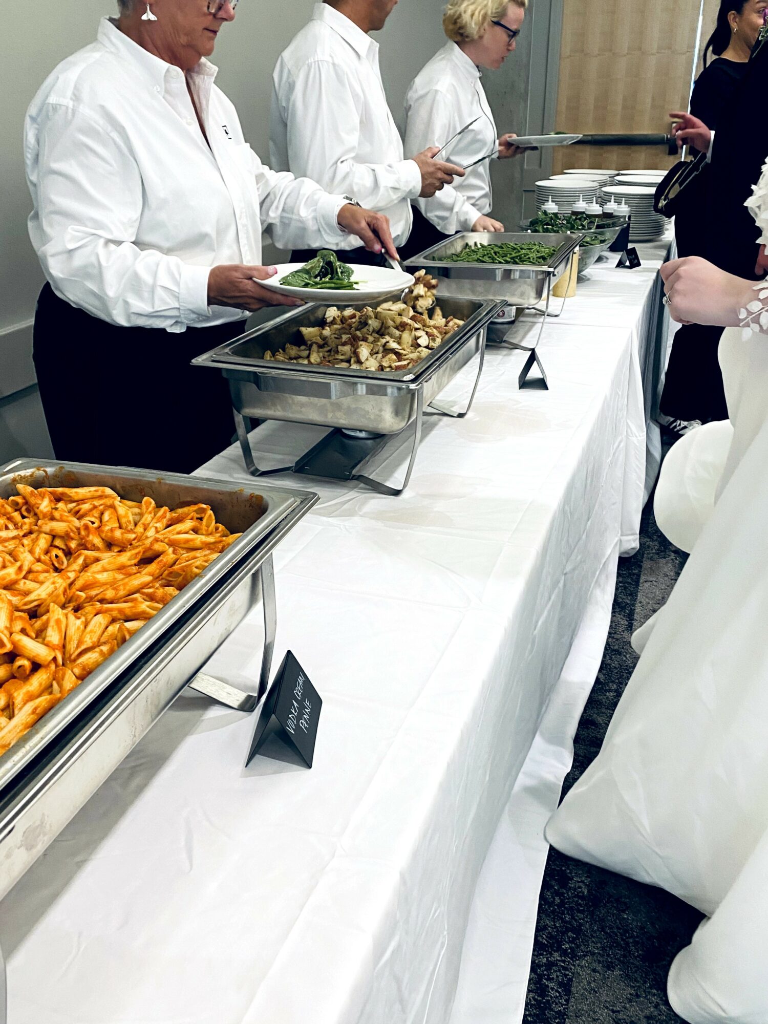 People serving food at a buffet with trays of pasta, vegetables, and other dishes on a white tablecloth.