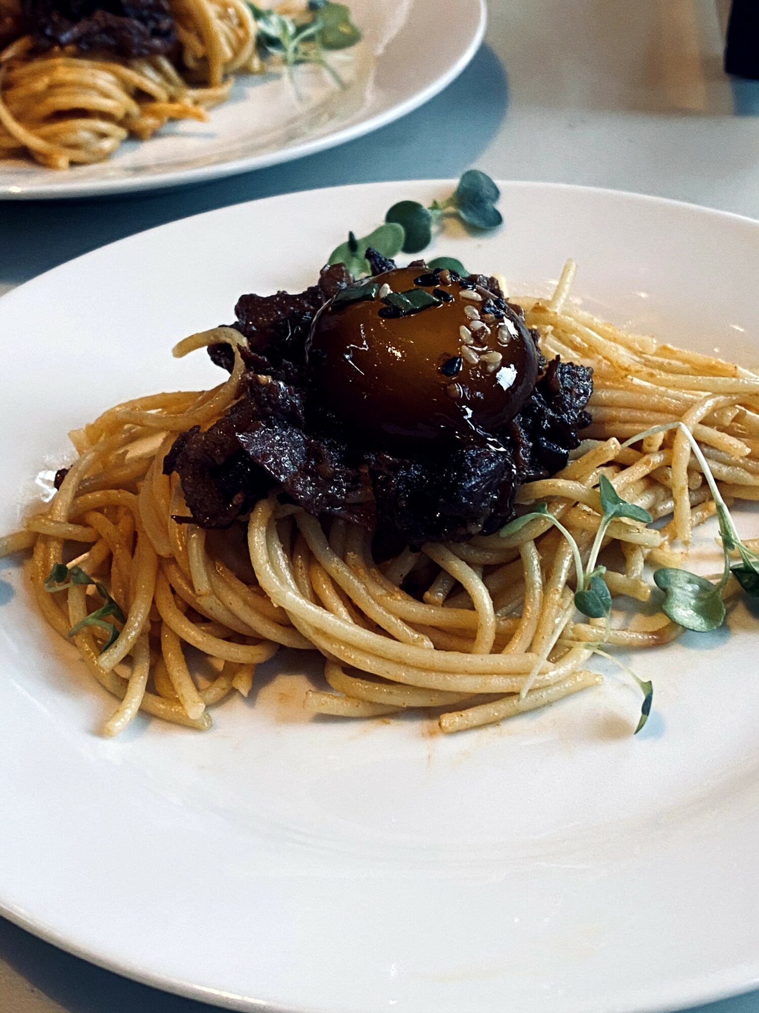 Spaghetti pasta topped with a dark sauce, a small vegetable, and microgreens on a white plate.