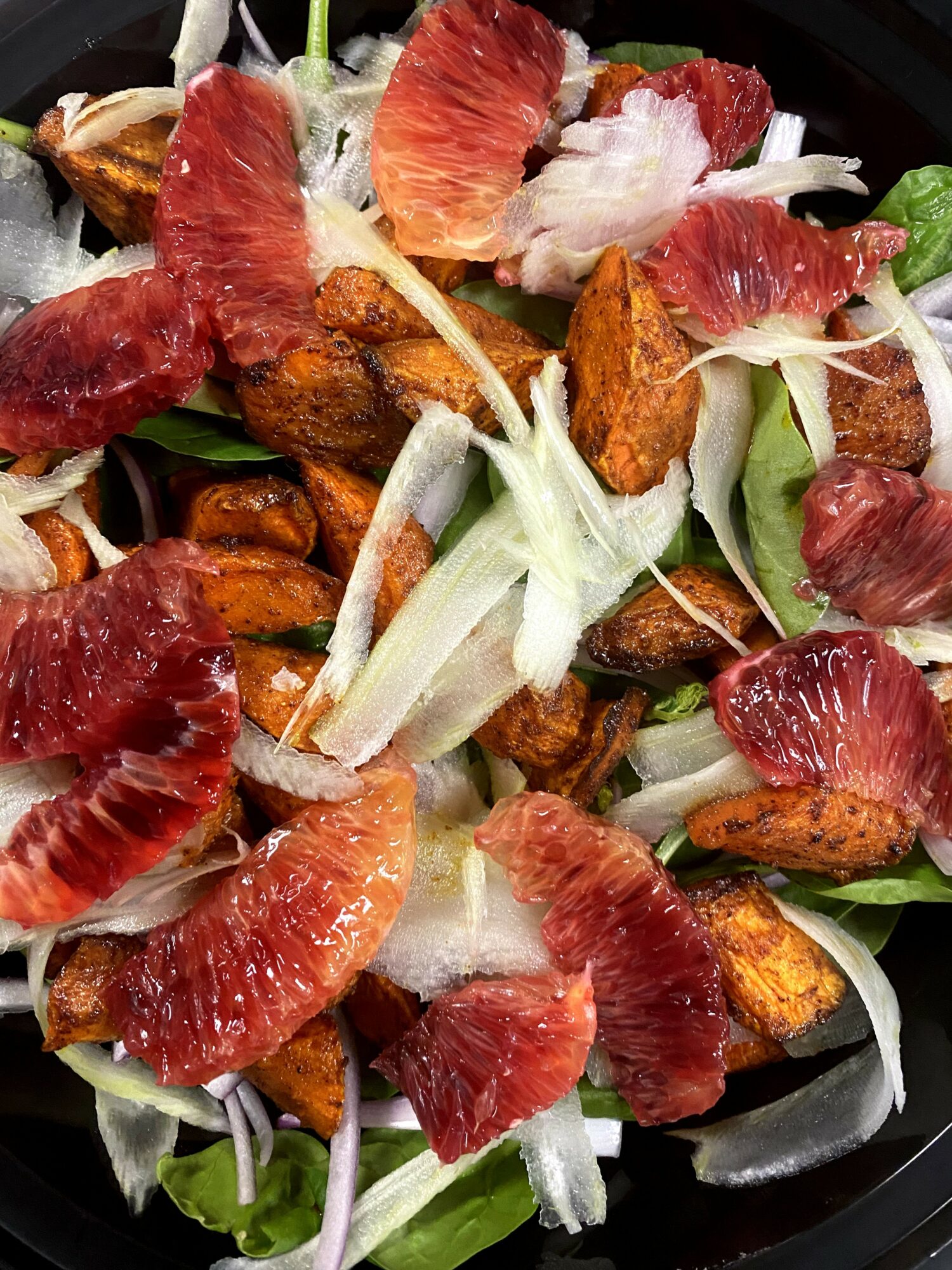 Salad with grapefruit segments, fried chicken pieces, and sliced onions on a black plate.