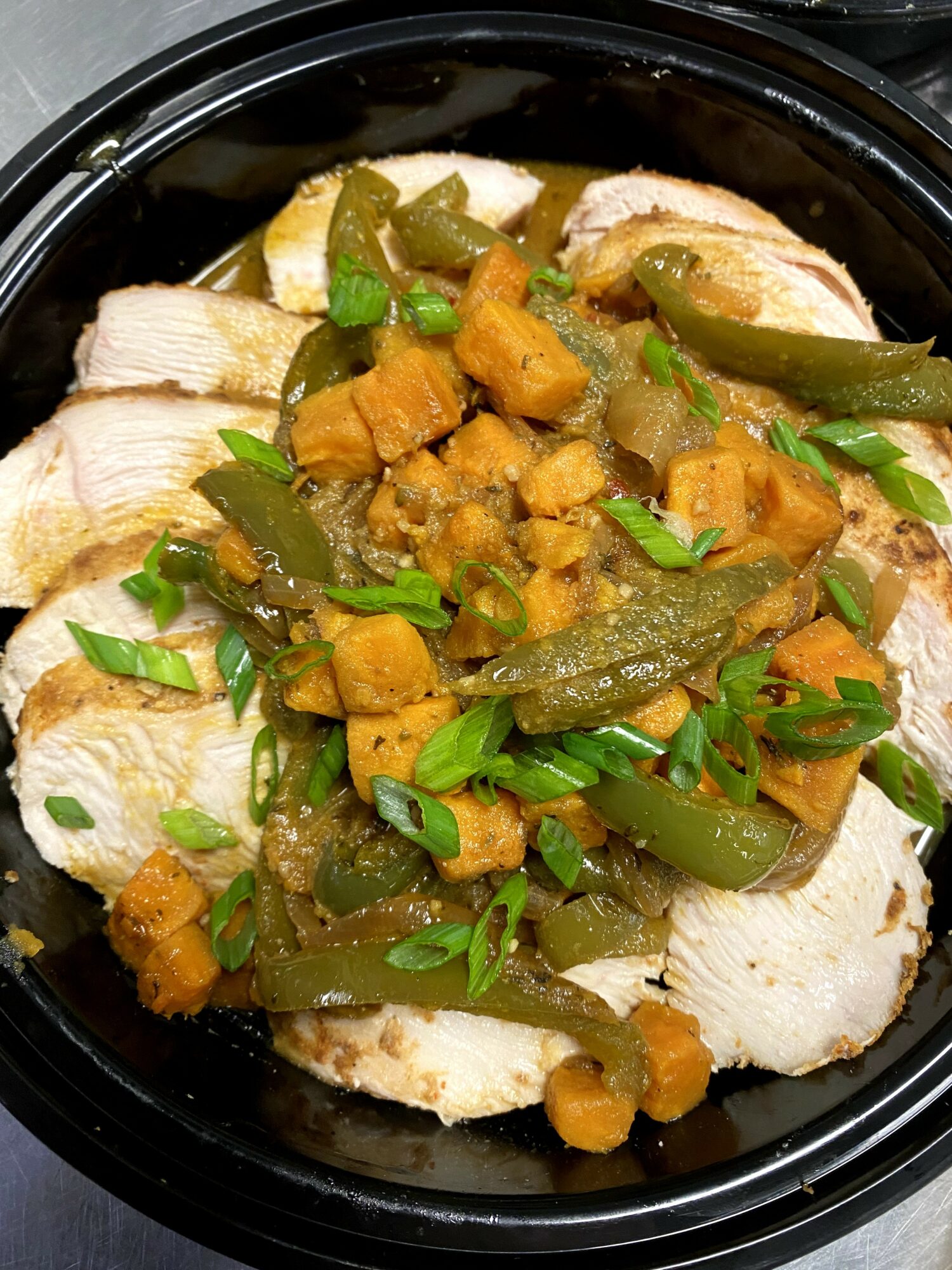 Sliced cooked chicken topped with diced vegetables and green herbs in a black container.