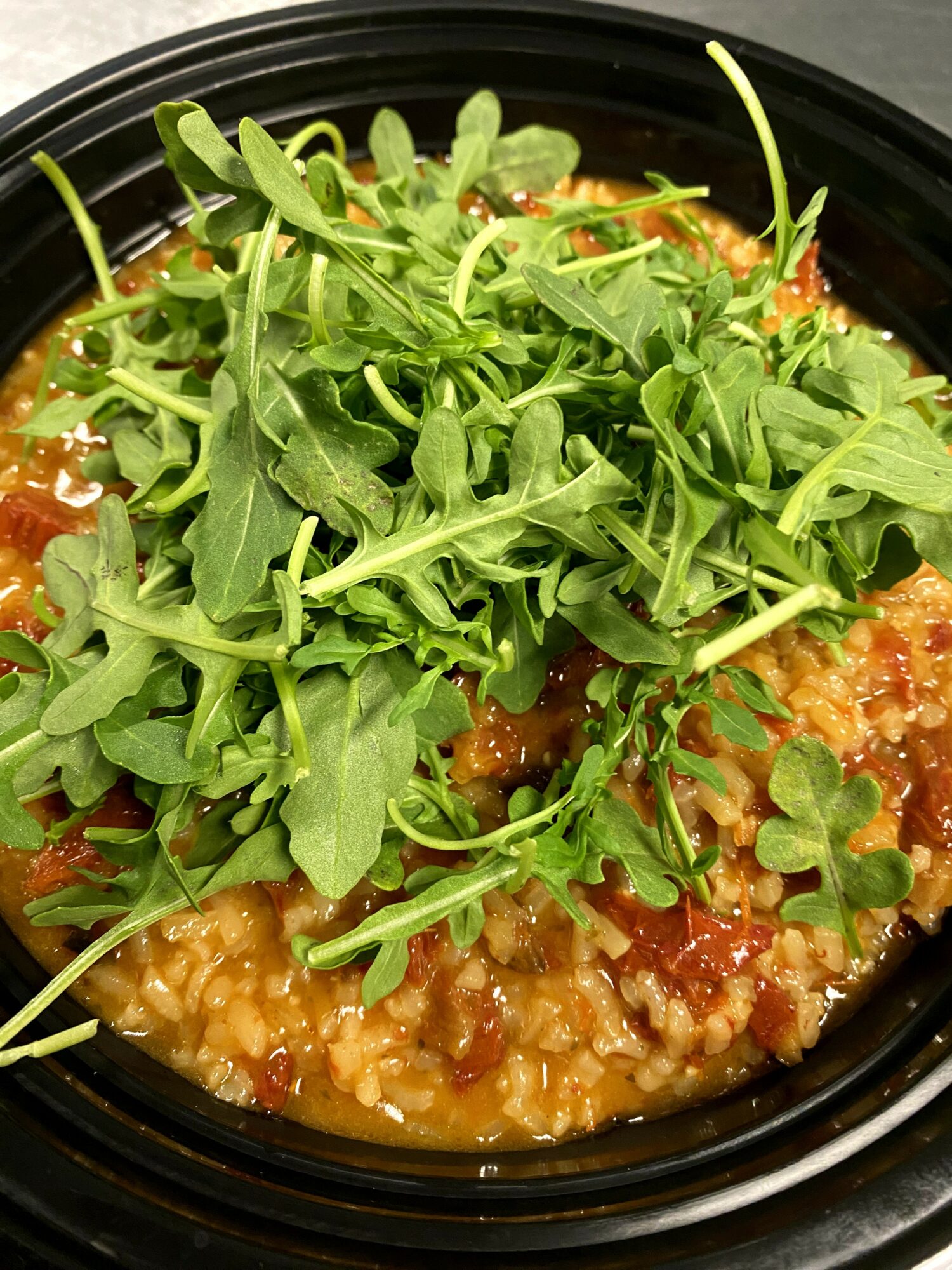 Dish with cooked grains, topped with fresh green arugula leaves in a black container.
