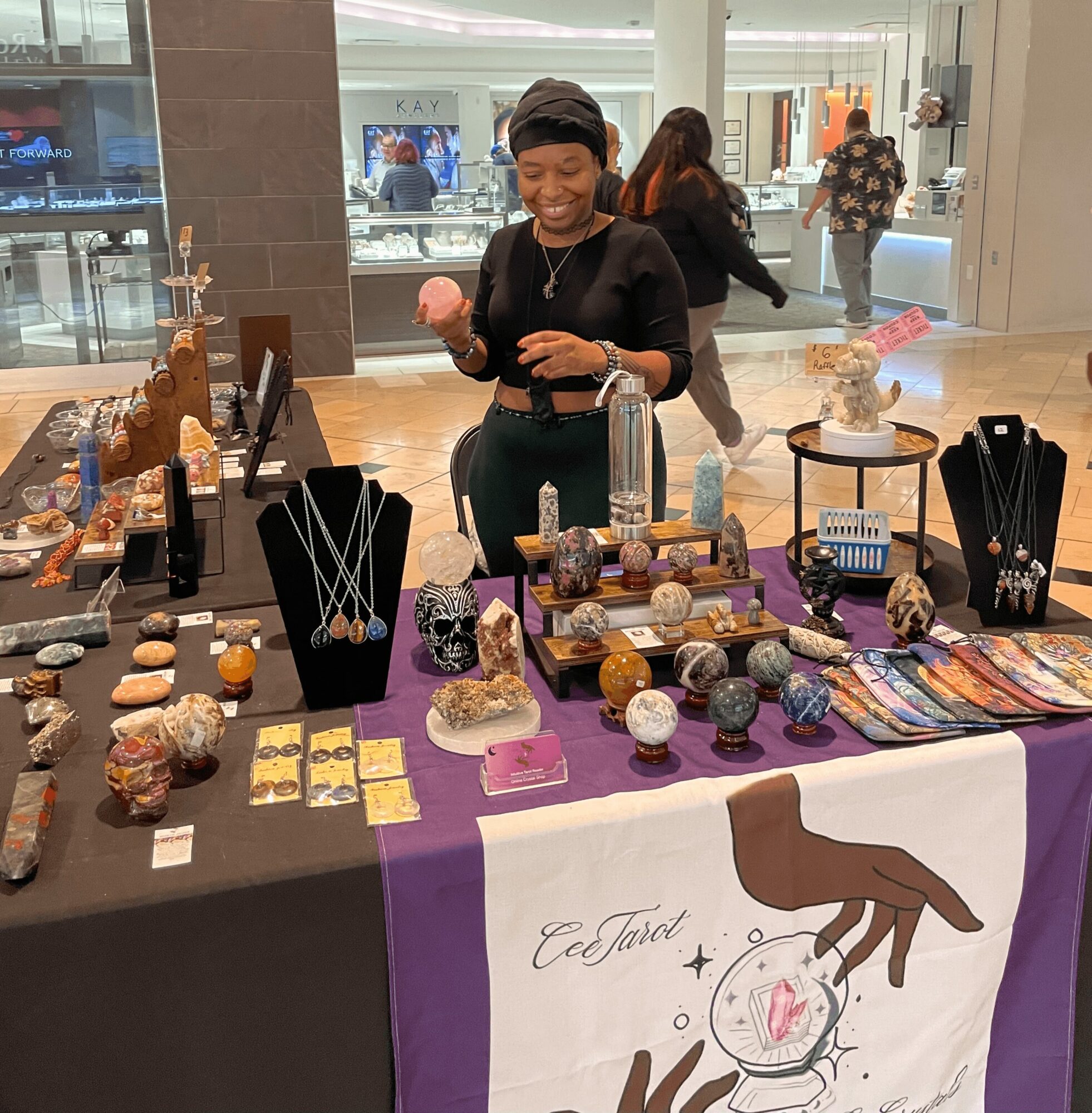 Smiling woman at jewelry and craft display in shopping mall, holding pink item, with other shoppers in background.