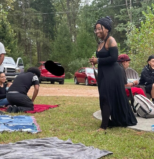 Woman in black dress standing outdoors with people sitting on blankets and cars in background.