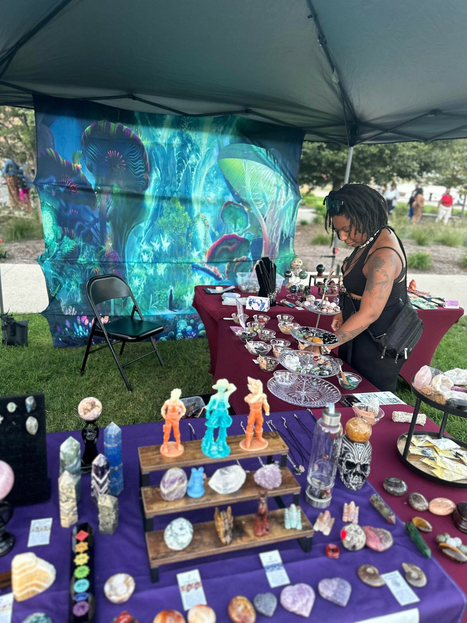 Woman browsing jewelry display at outdoor market with colorful artwork backdrop, tables with various jewelry items, and a tent overhead.