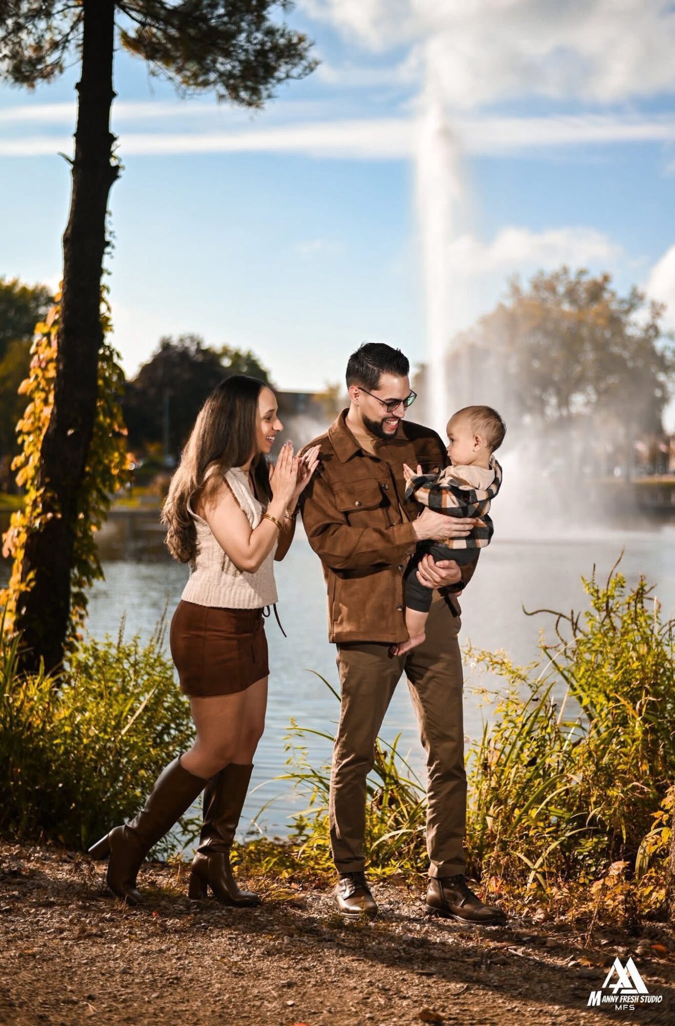 Family of three by a lake with a fountain, trees, and blue sky, enjoying a moment together outdoors.
