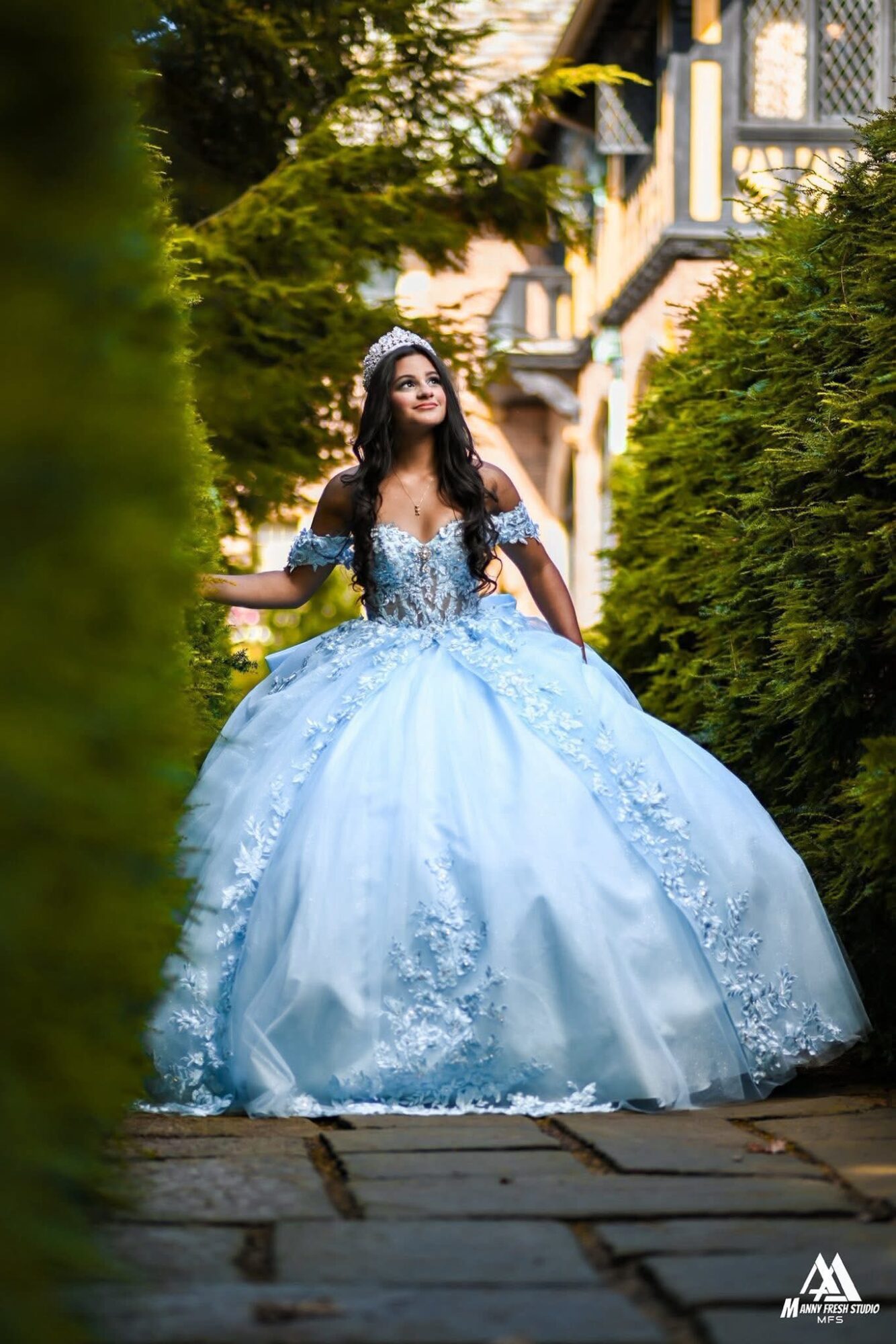 Young woman in a light blue ball gown standing on a stone pathway surrounded by greenery, smiling, with a building in the background.