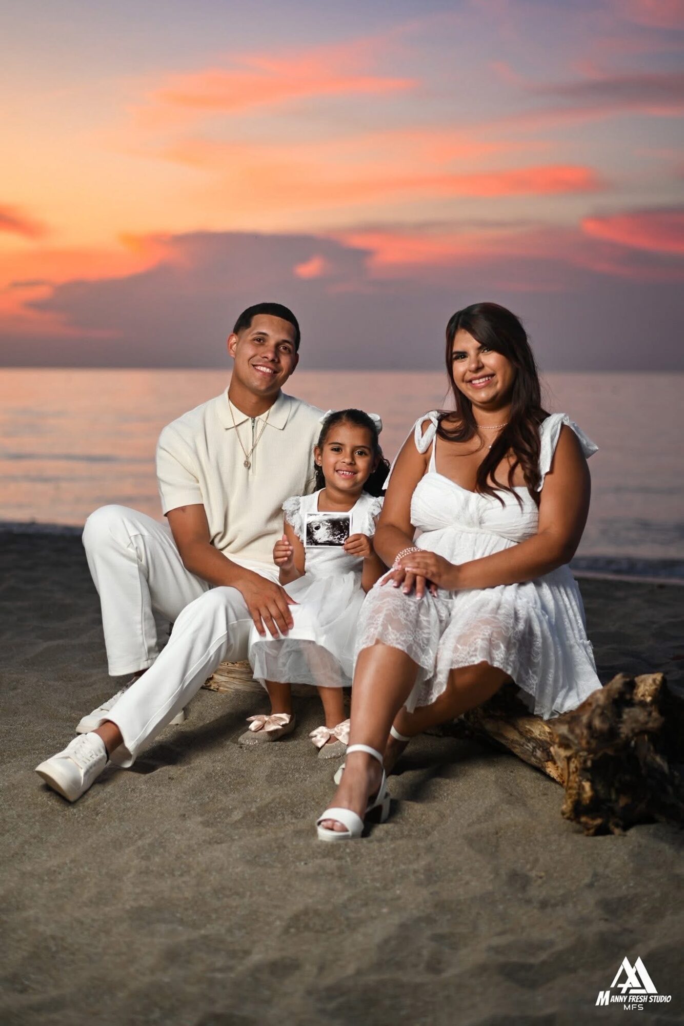 Family of three sitting on the beach at sunset, smiling, with colorful sky and ocean in background.