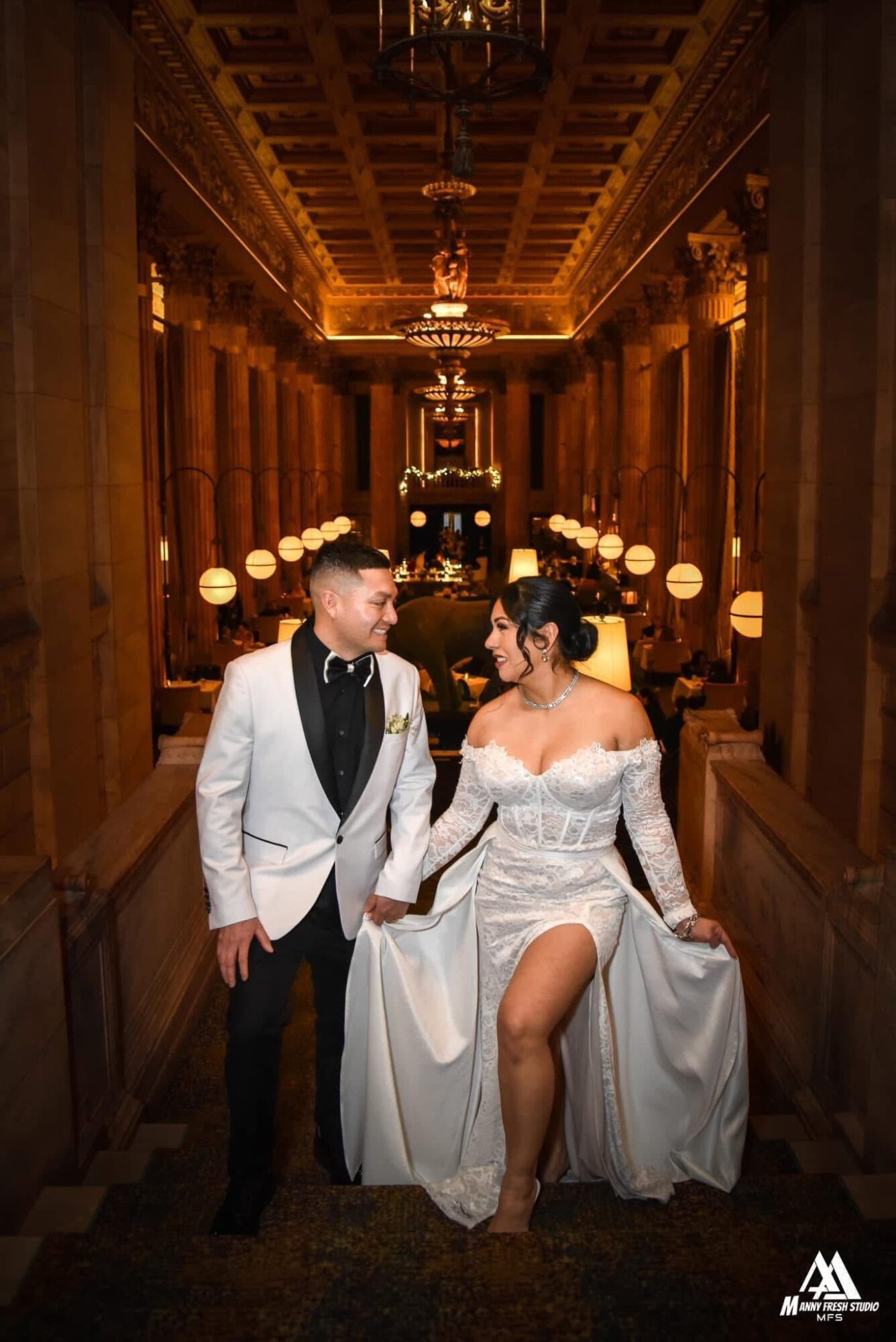A couple in wedding attire sitting in an ornate, dimly lit hall with chandeliers and wooden paneling.