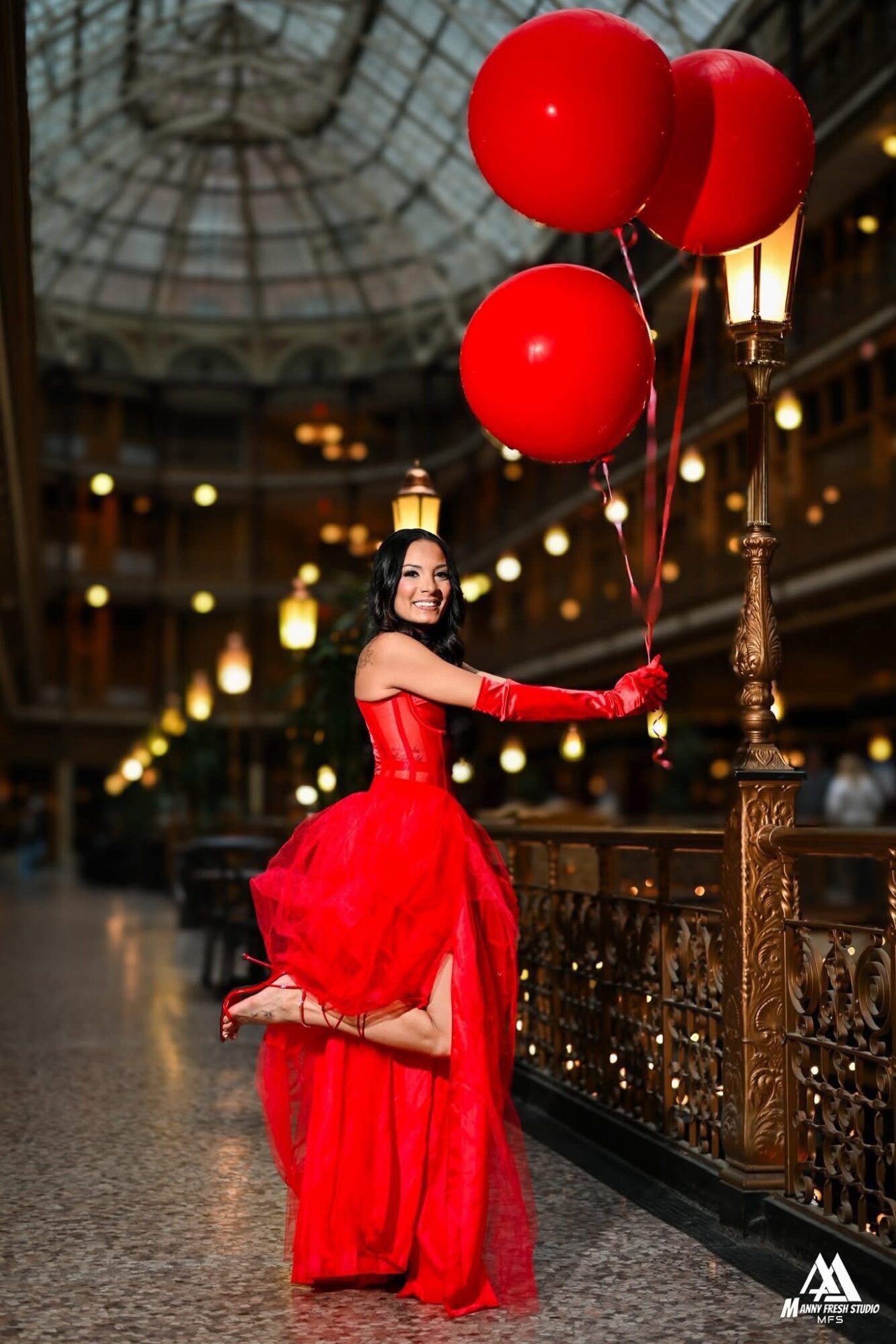 Woman in red dress holding three red balloons inside a grand, ornate building with a glass ceiling and warm lighting.