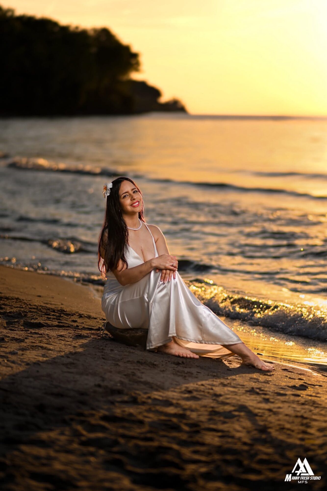 Woman in white dress sitting on beach at sunset, smiling, with water and trees in background.