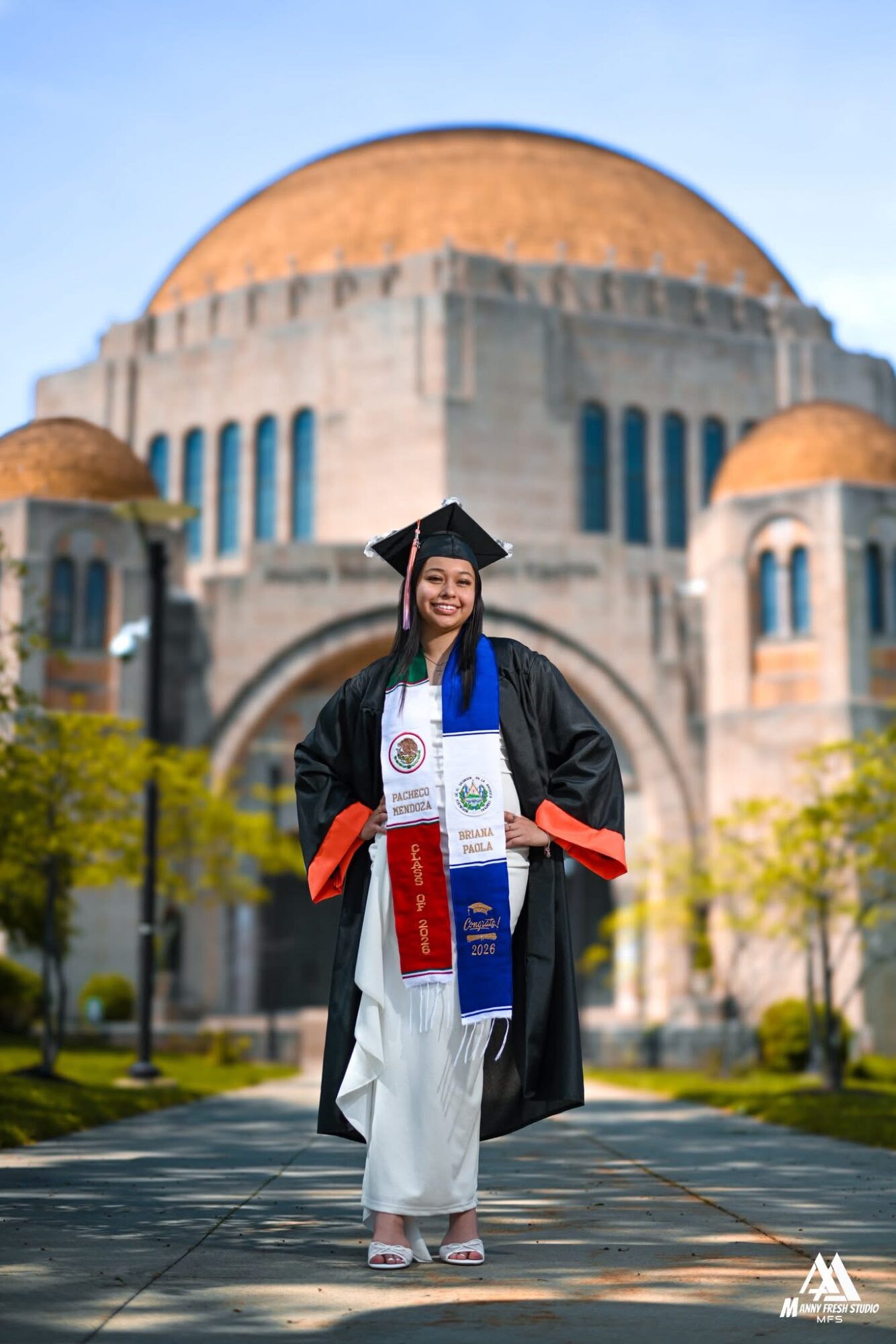 Young woman in graduation gown and cap standing outdoors in front of a large domed building, smiling, with trees and a pathway.