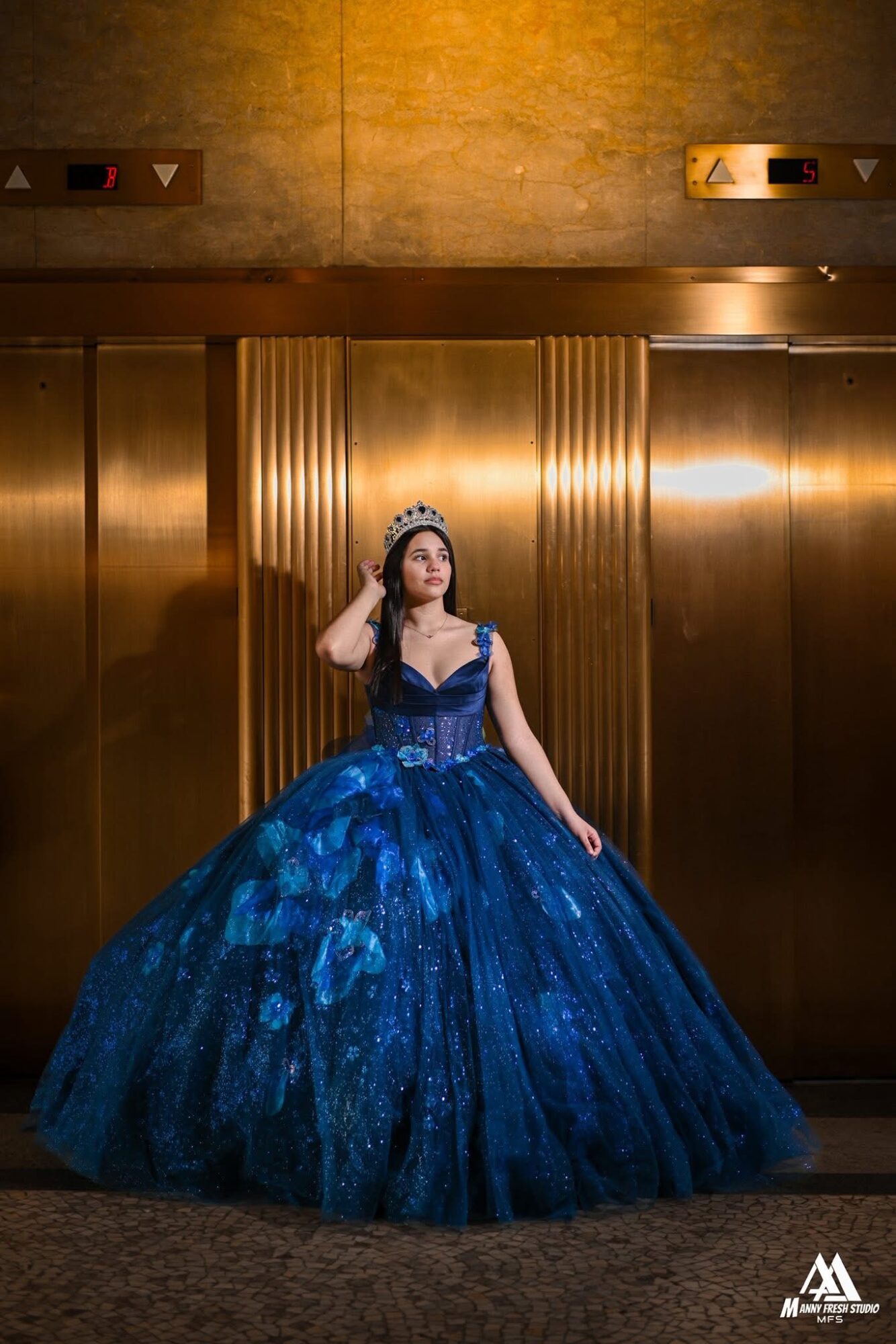 Woman in a blue gown and tiara stands in front of gold-colored elevator doors, with a textured wall above.