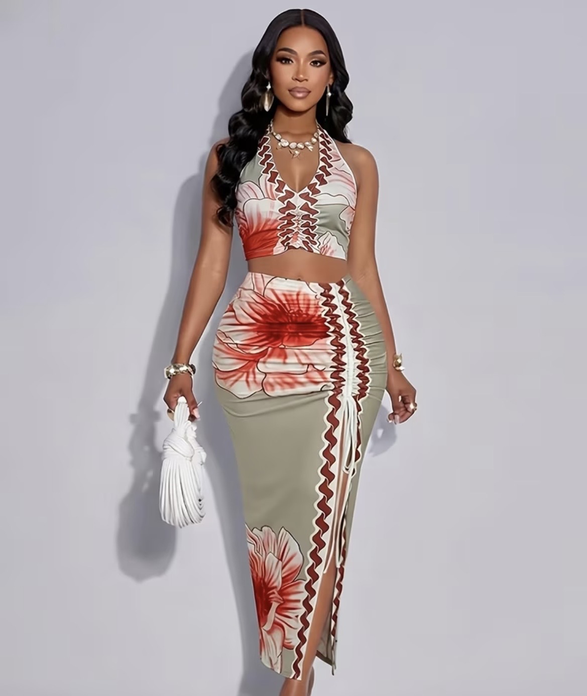Woman in floral crop top and skirt holding a white bag, standing against a plain background.