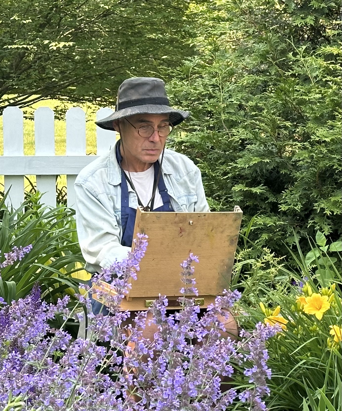 Person wearing a hat and glasses painting outdoors among purple and yellow flowers, with green trees and a white fence in background.