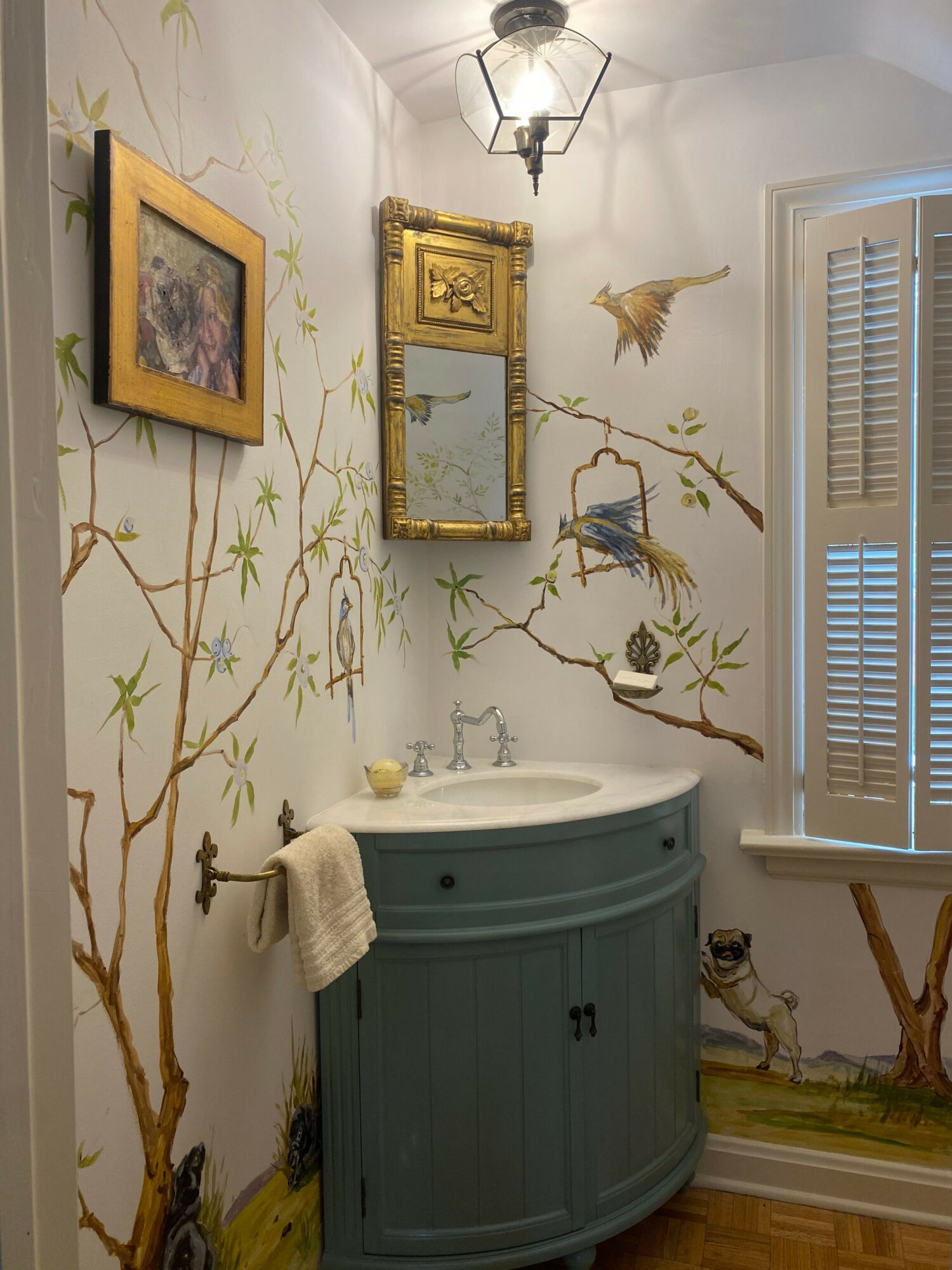 Wall with painted branches and birds, a clock, a framed picture, and a lamp, with a round gray object and a towel.
