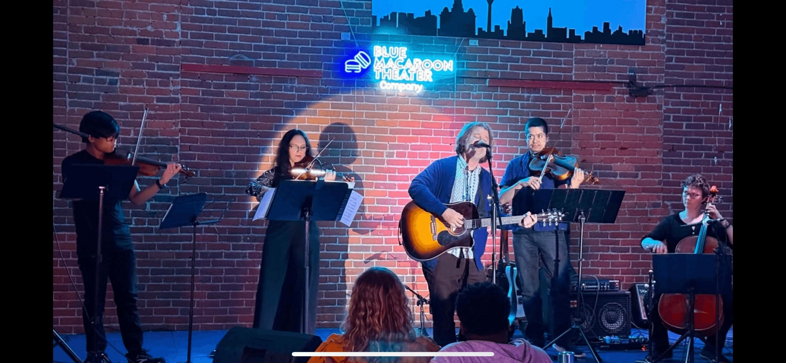 Group of five musicians performing on stage with brick wall background and neon sign, one playing guitar, others singing or playing instruments.