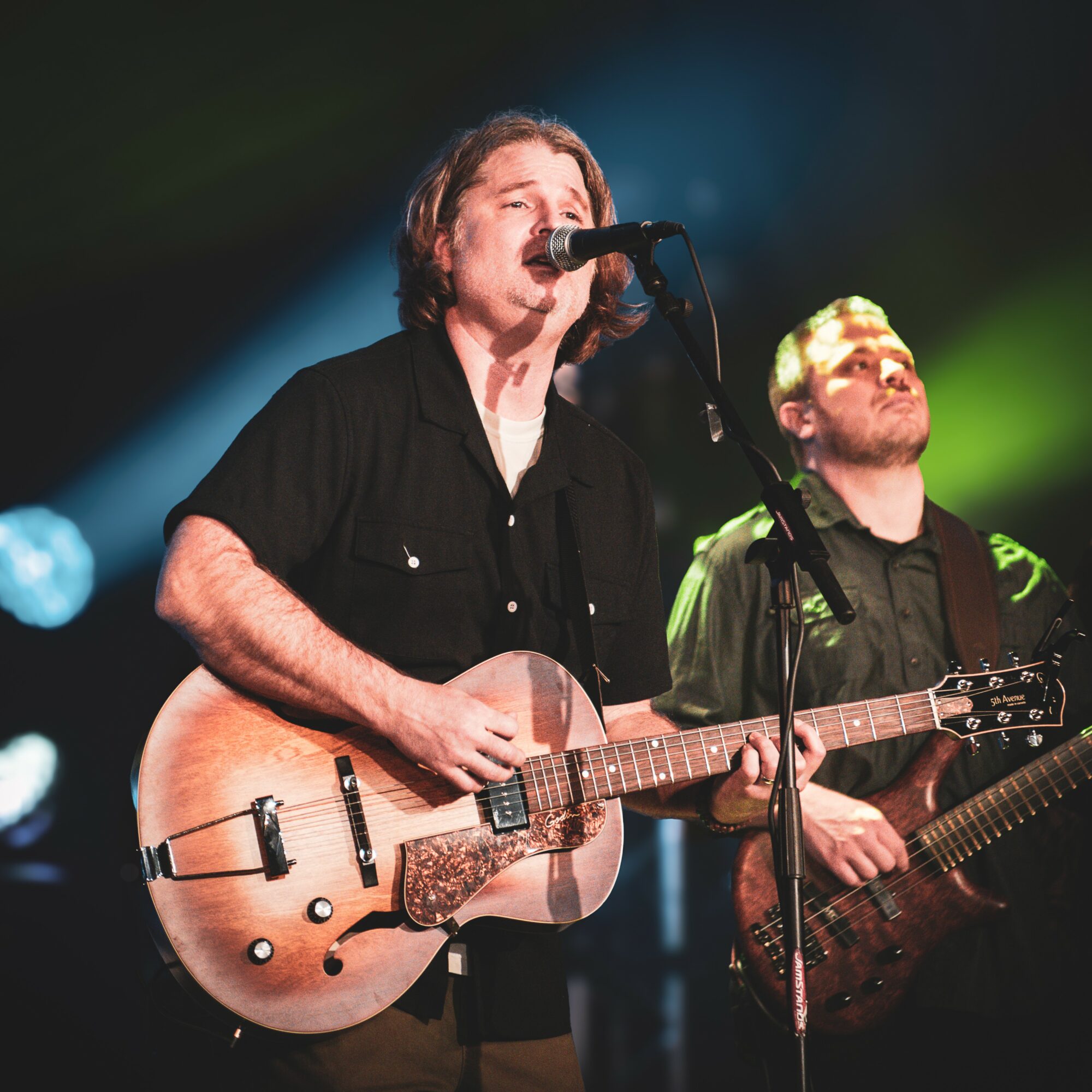 Two musicians performing on stage, one singing into a microphone and playing an acoustic guitar, the other playing an electric bass guitar, with colorful stage lighting.
