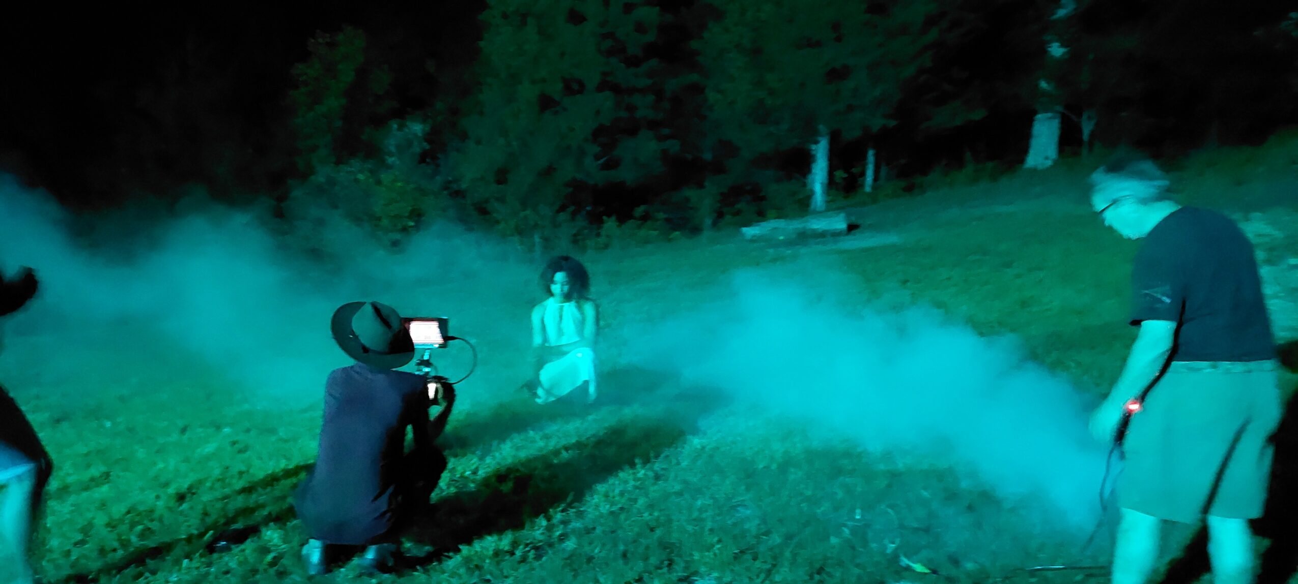 Group of people outdoors at night, some wearing virtual reality headsets, surrounded by fog or smoke, with trees in the background.