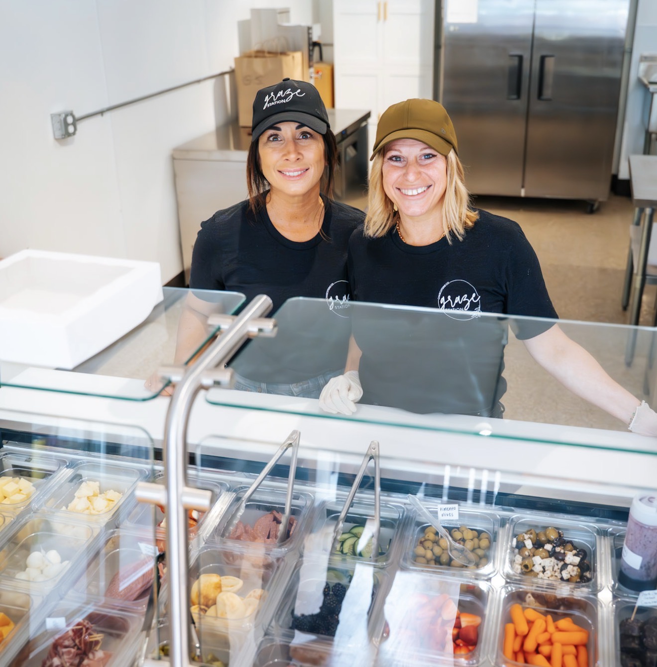 Two smiling women behind a glass display counter in a kitchen or bakery setting, wearing black shirts and hats.
