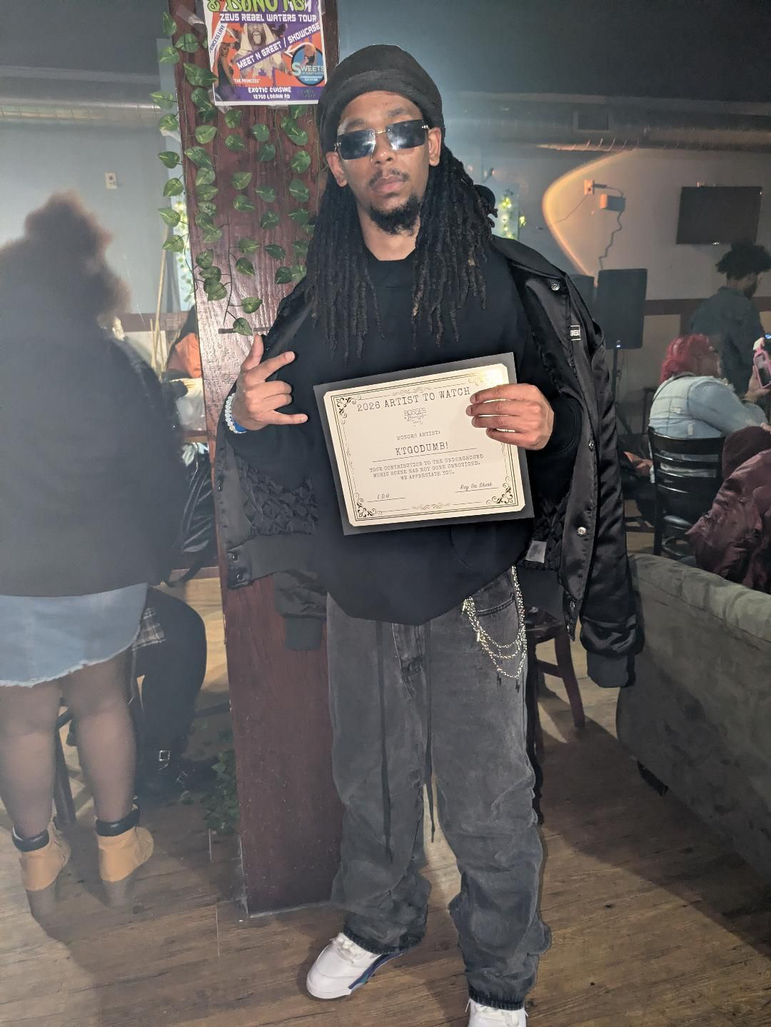 Person with long dreadlocks, sunglasses, black jacket, holding a certificate, standing outdoors at night with people in background.