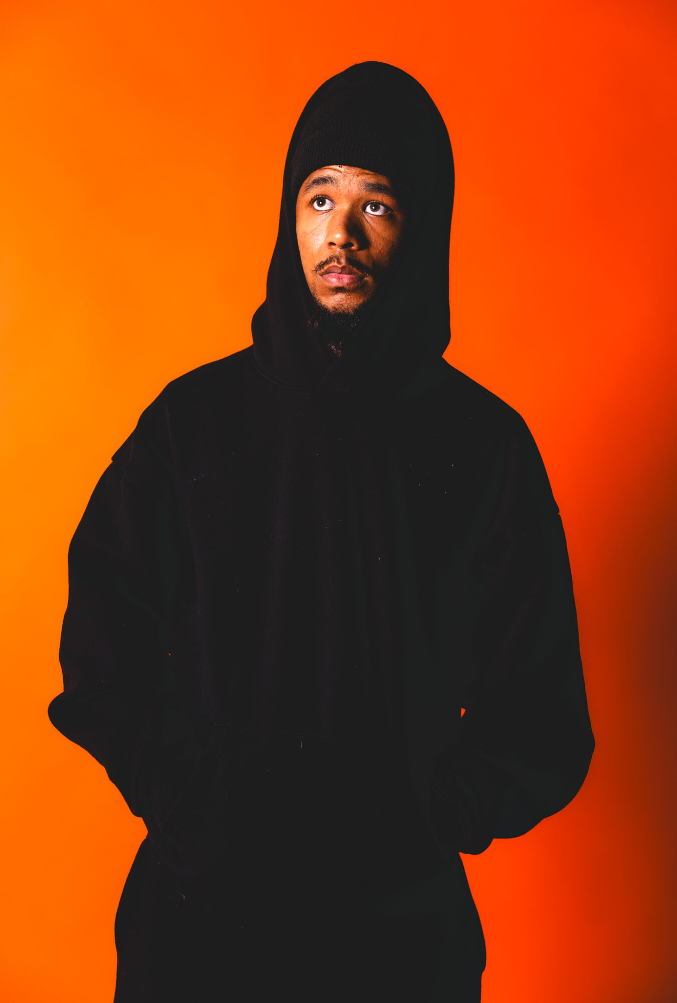 Person wearing a black hoodie and beanie against an orange background, looking to the side.