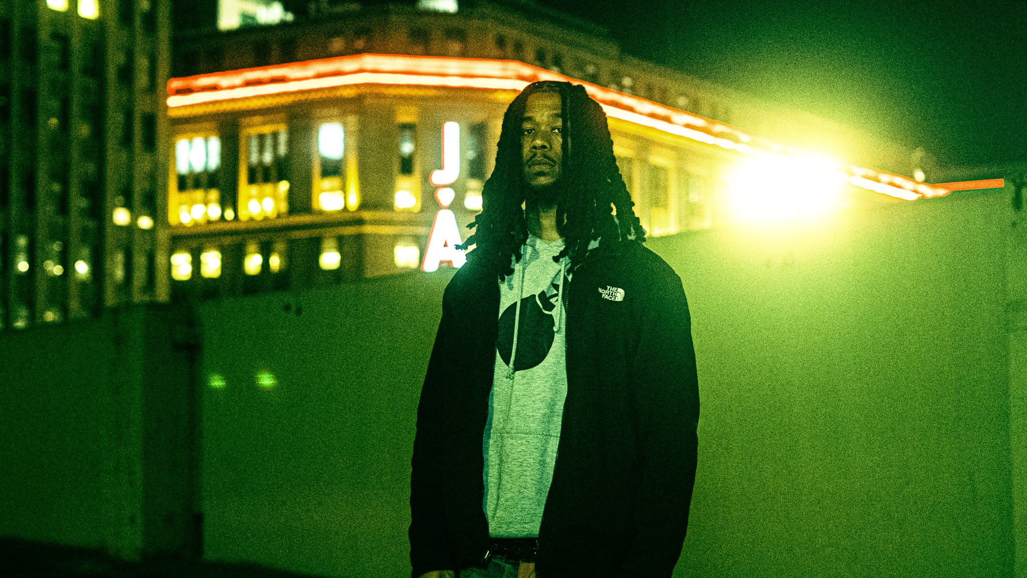 Person with long hair standing outdoors at night with city lights in background, illuminated by bright greenish light.