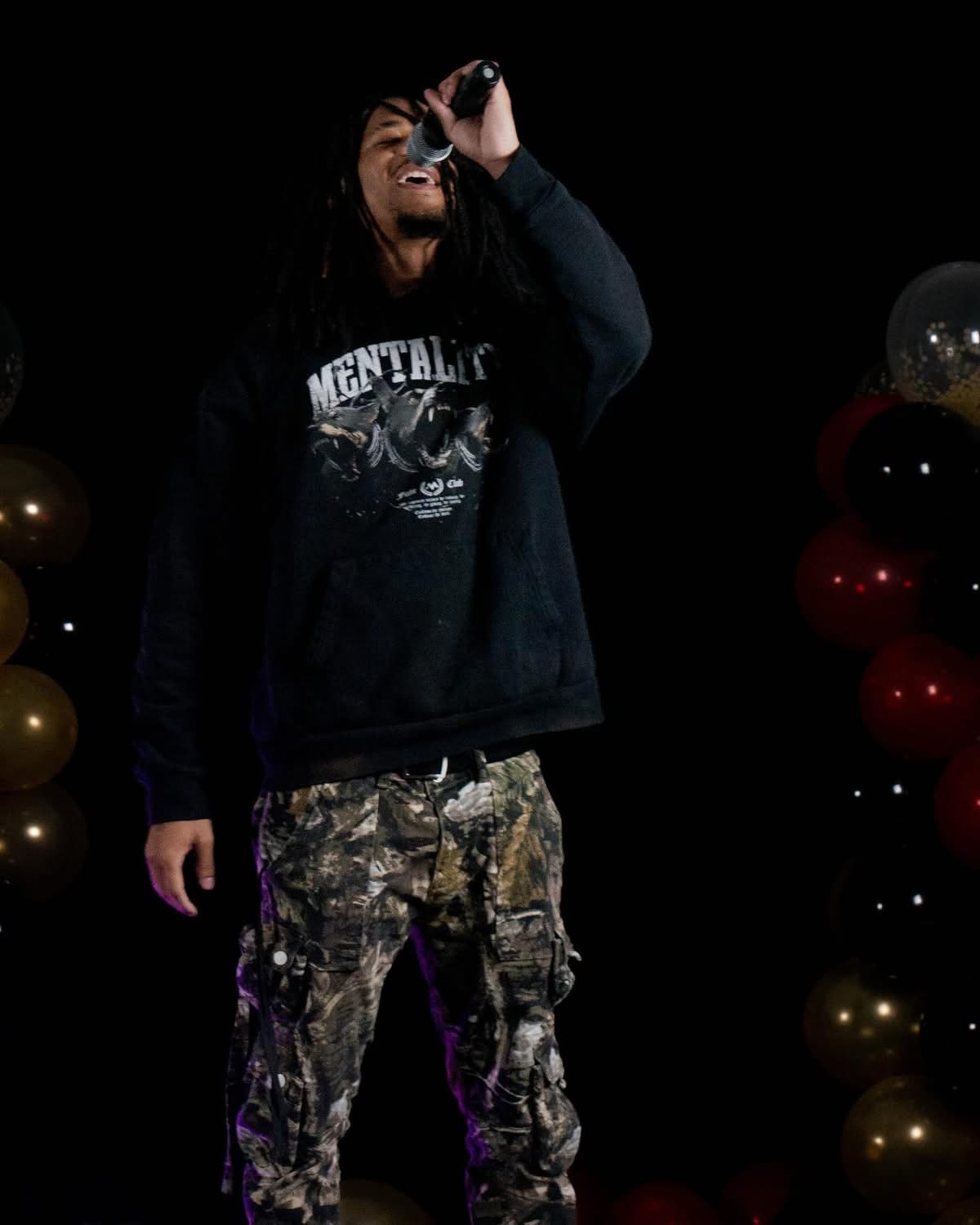 Person standing in front of balloons, wearing a black hoodie and camouflage pants, holding a microphone to their mouth.