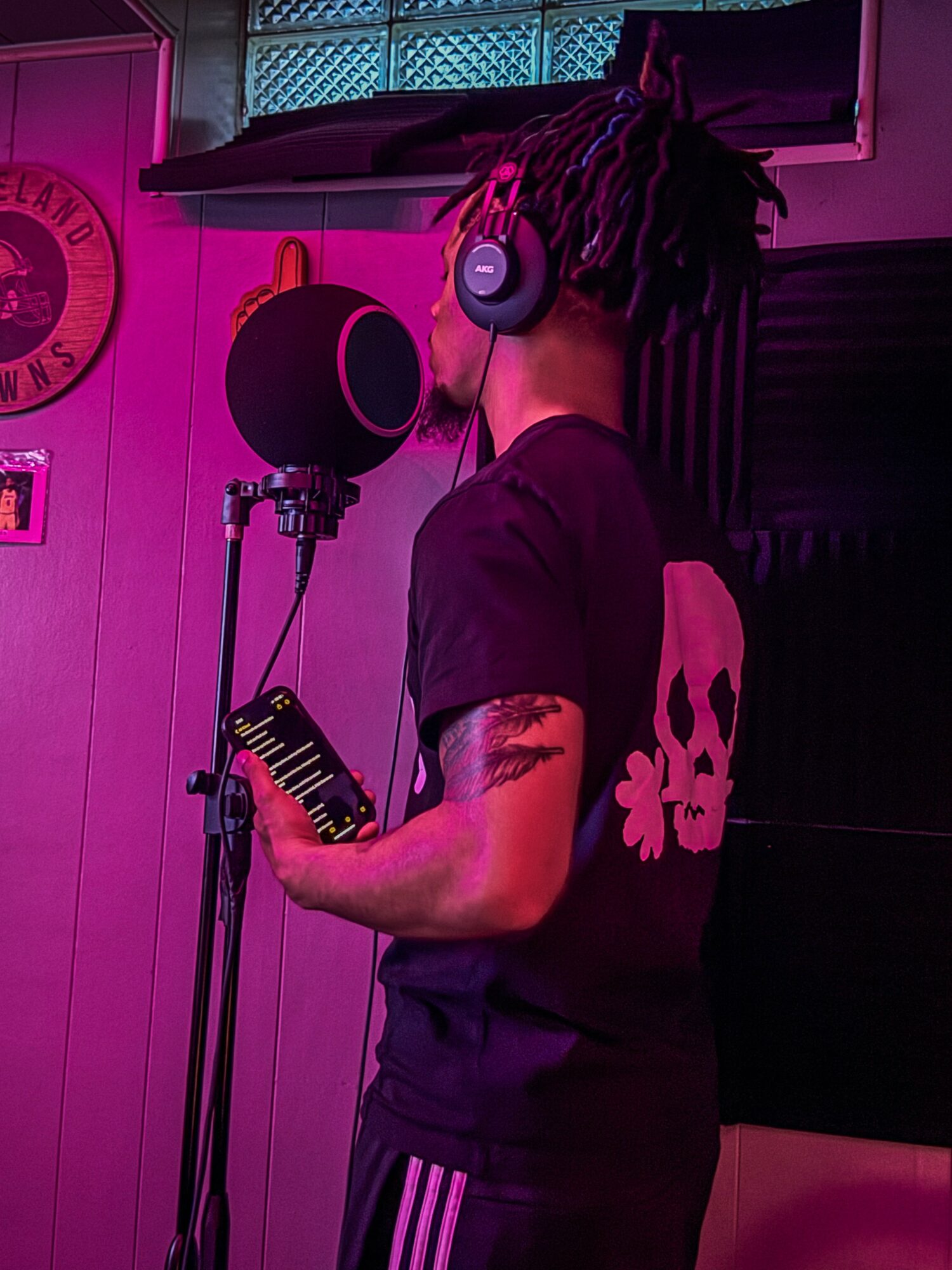Person with dreadlocks wearing headphones, standing in front of a microphone in a recording studio, holding a phone.
