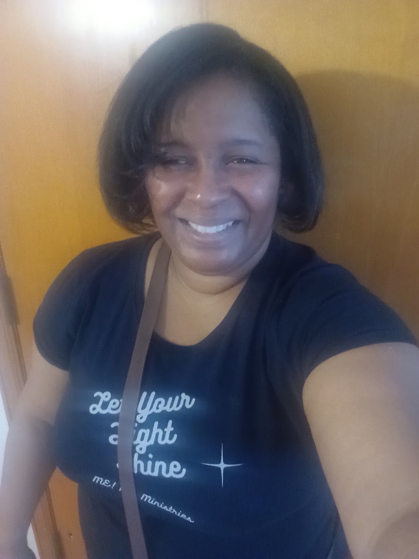 Smiling woman with shoulder-length dark hair taking a selfie indoors, wearing a black T-shirt and carrying a bag.