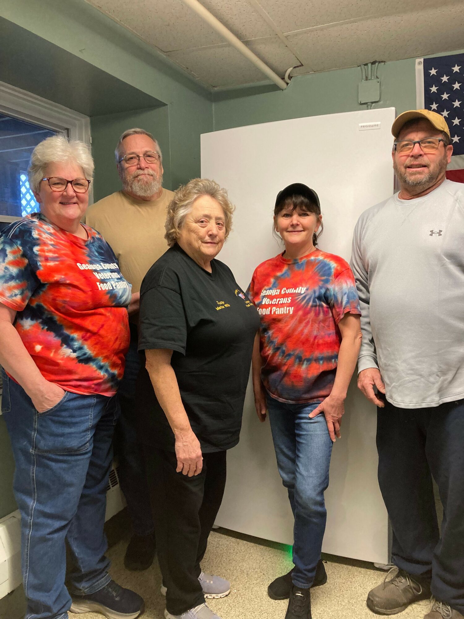 Four people standing indoors, two women and two men, with a white wall and an American flag in the background.