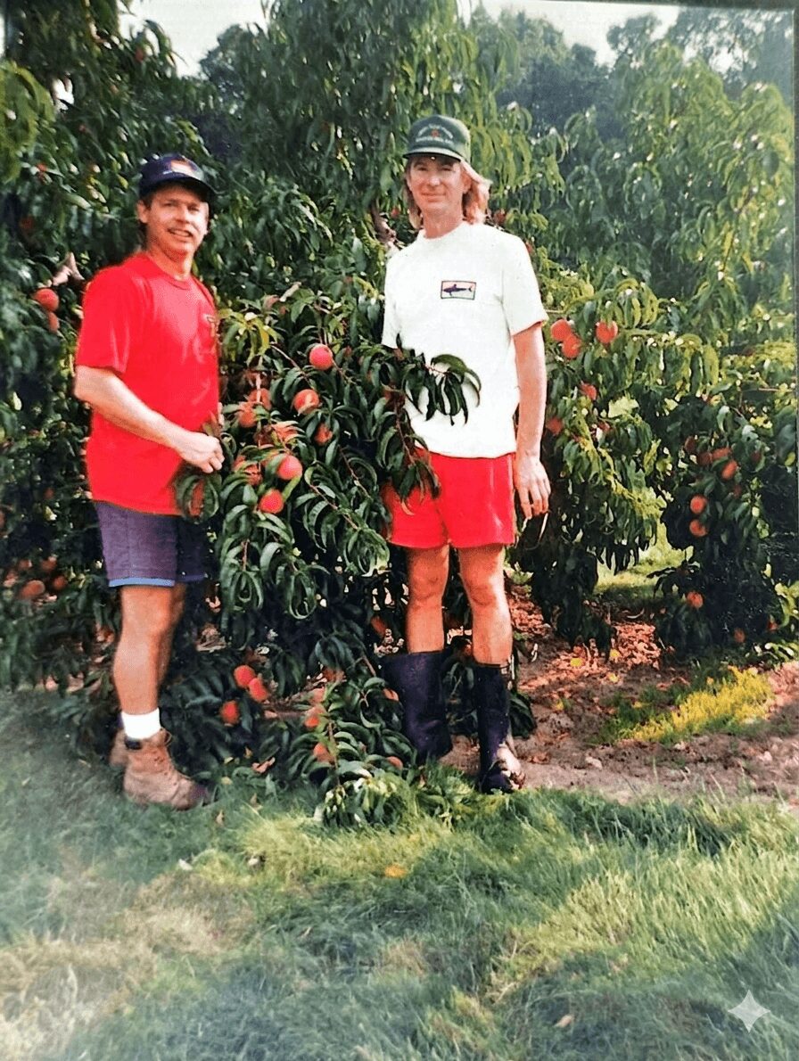 Two people stand outdoors among peach trees, one in a red shirt and shorts, the other in a white shirt and red shorts.