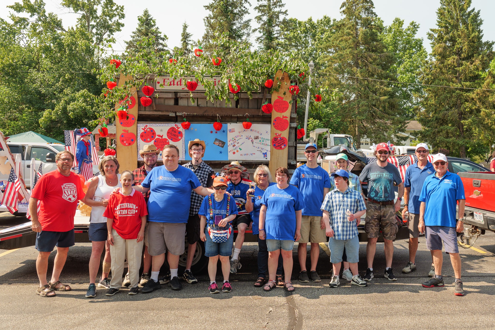 Group of people, including children and adults, standing outdoors in front of a decorated stage with trees in the background.
