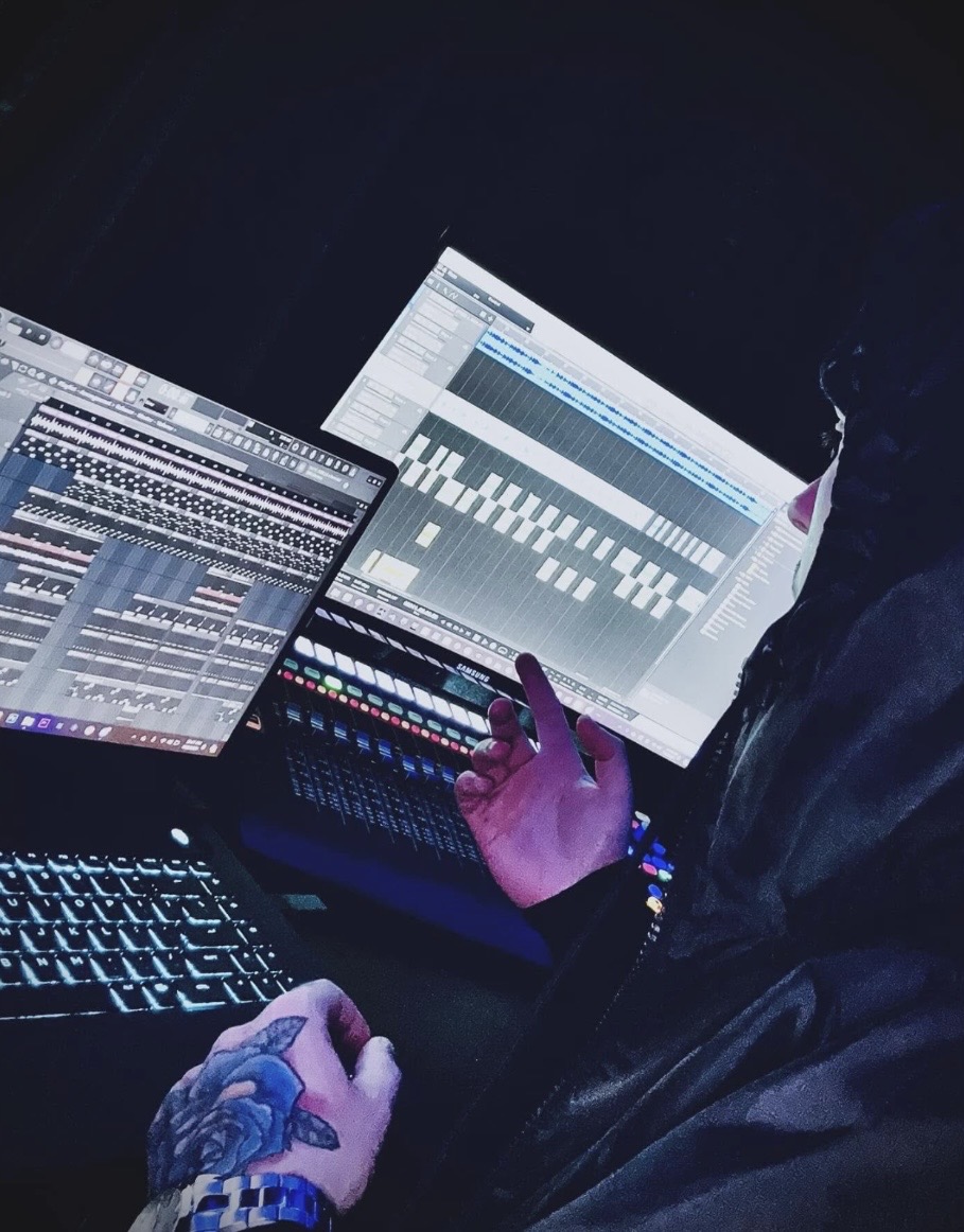 Person working with a laptop and two screens displaying music production software, wearing a dark hoodie and a tattooed hand.