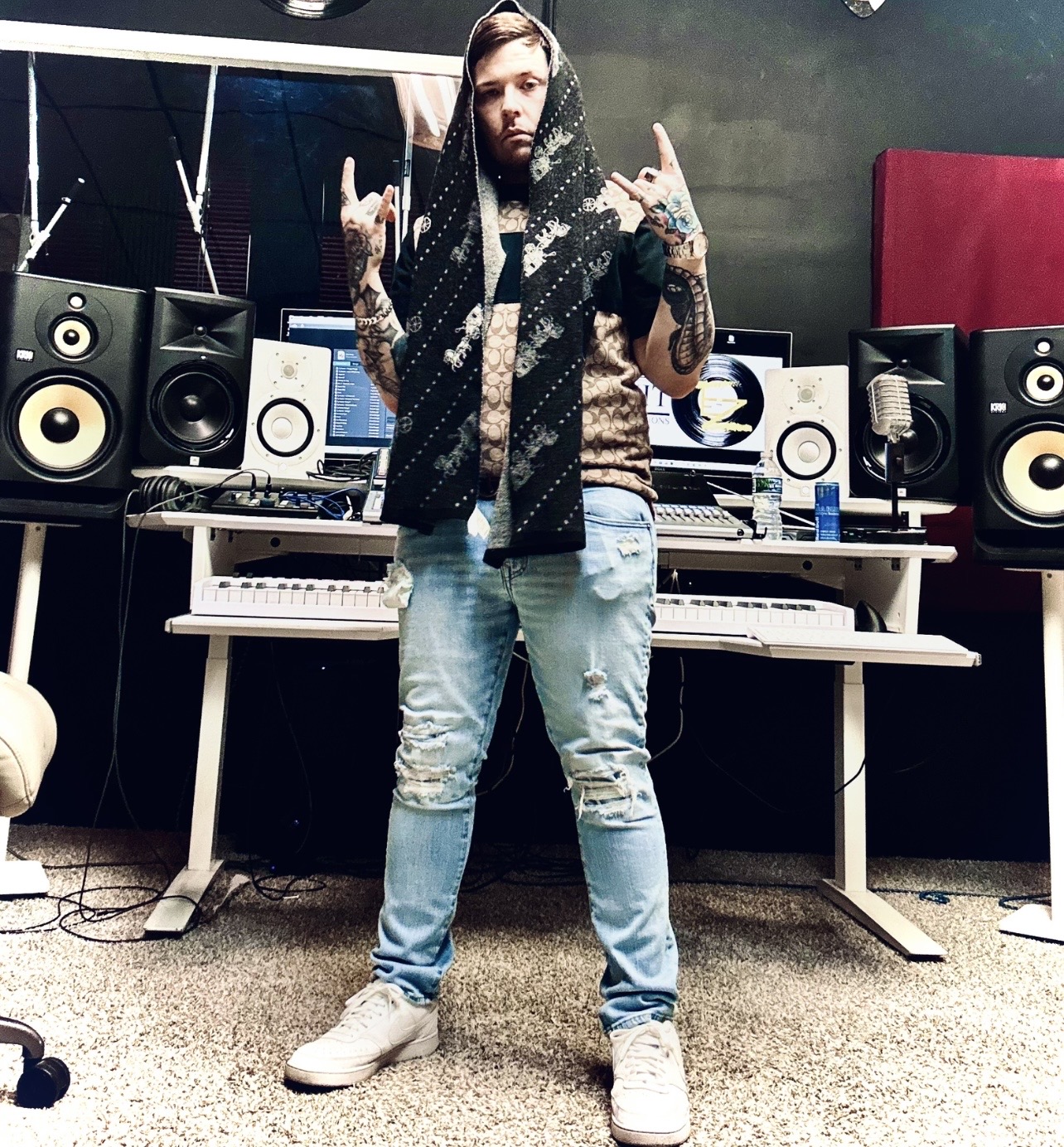 Person with long hair and tattoos standing in a music studio, making hand gestures, surrounded by speakers and music equipment.