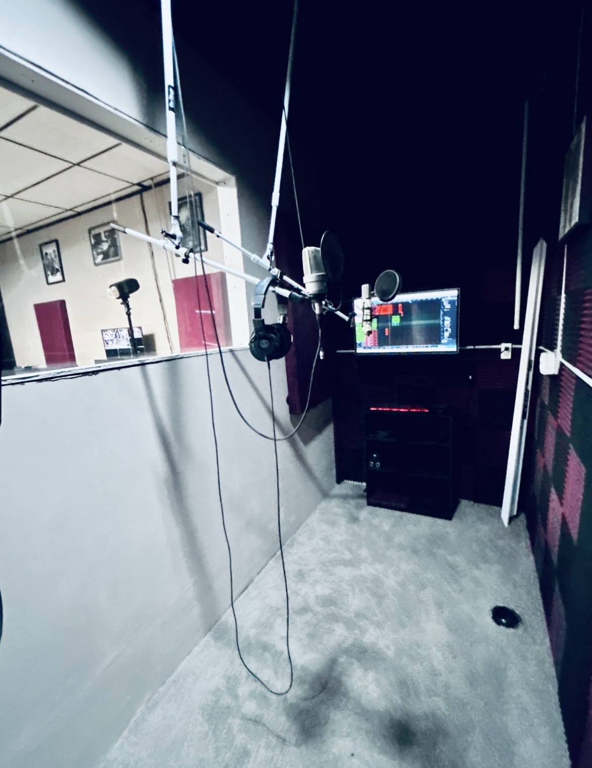 Recording studio with microphone, headphones, and audio equipment on a desk, viewed from the side, with a window into the control room.