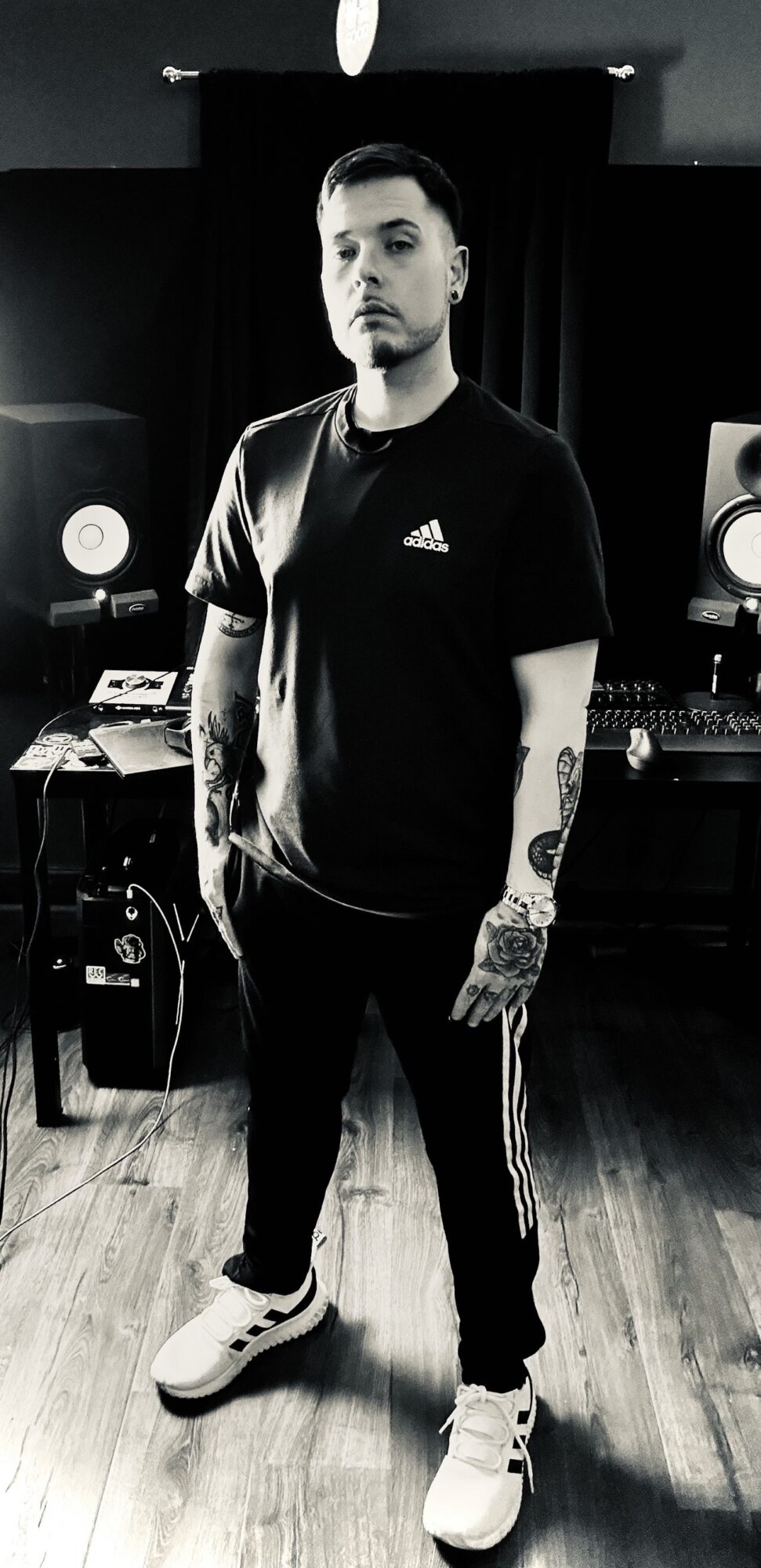 Young man with tattoos standing in a music studio, wearing a black Adidas t-shirt, black pants with white stripes, and white sneakers.