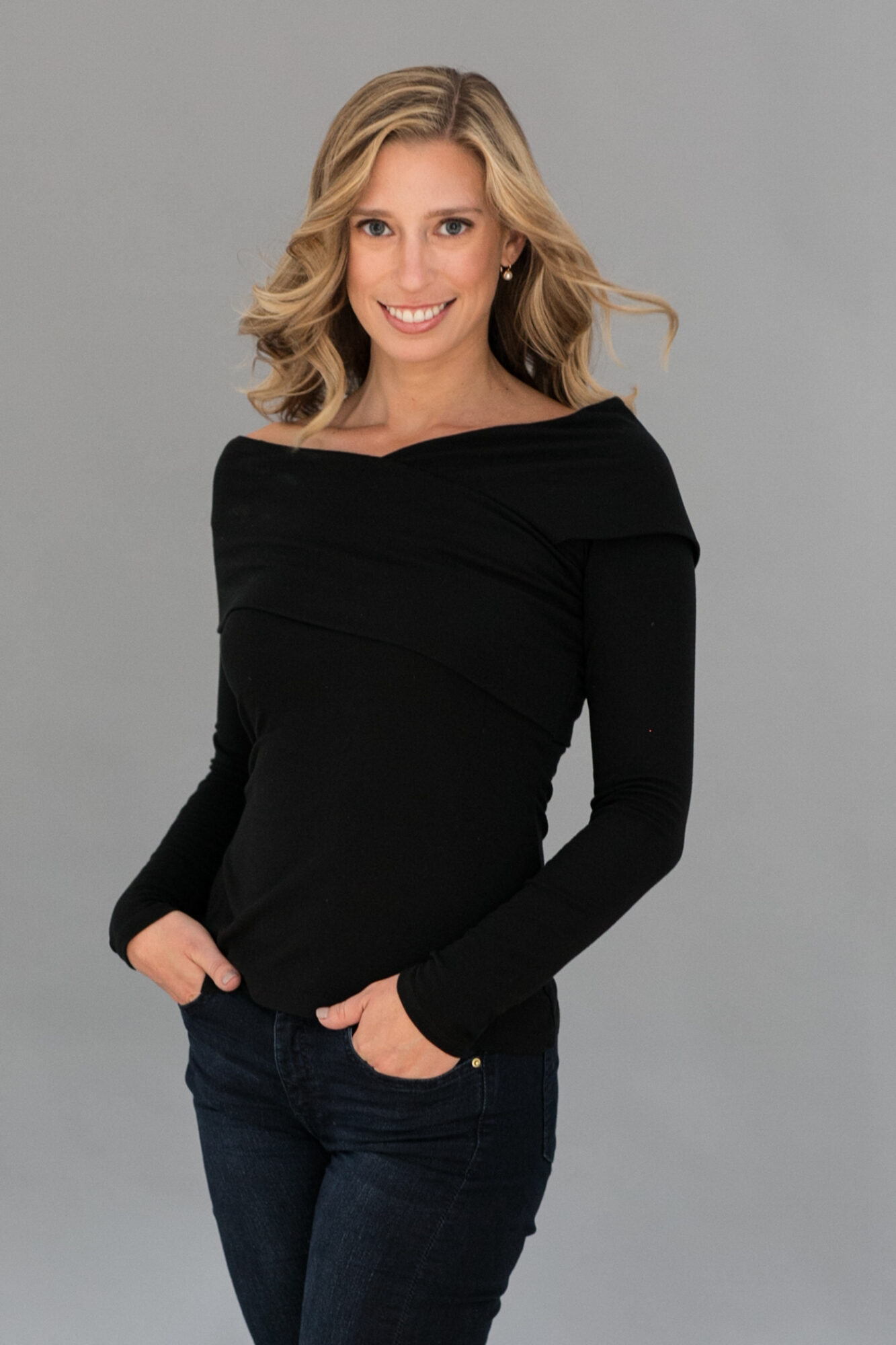 Woman with blonde hair smiling, wearing a black long-sleeve top and dark jeans, standing against a gray background.