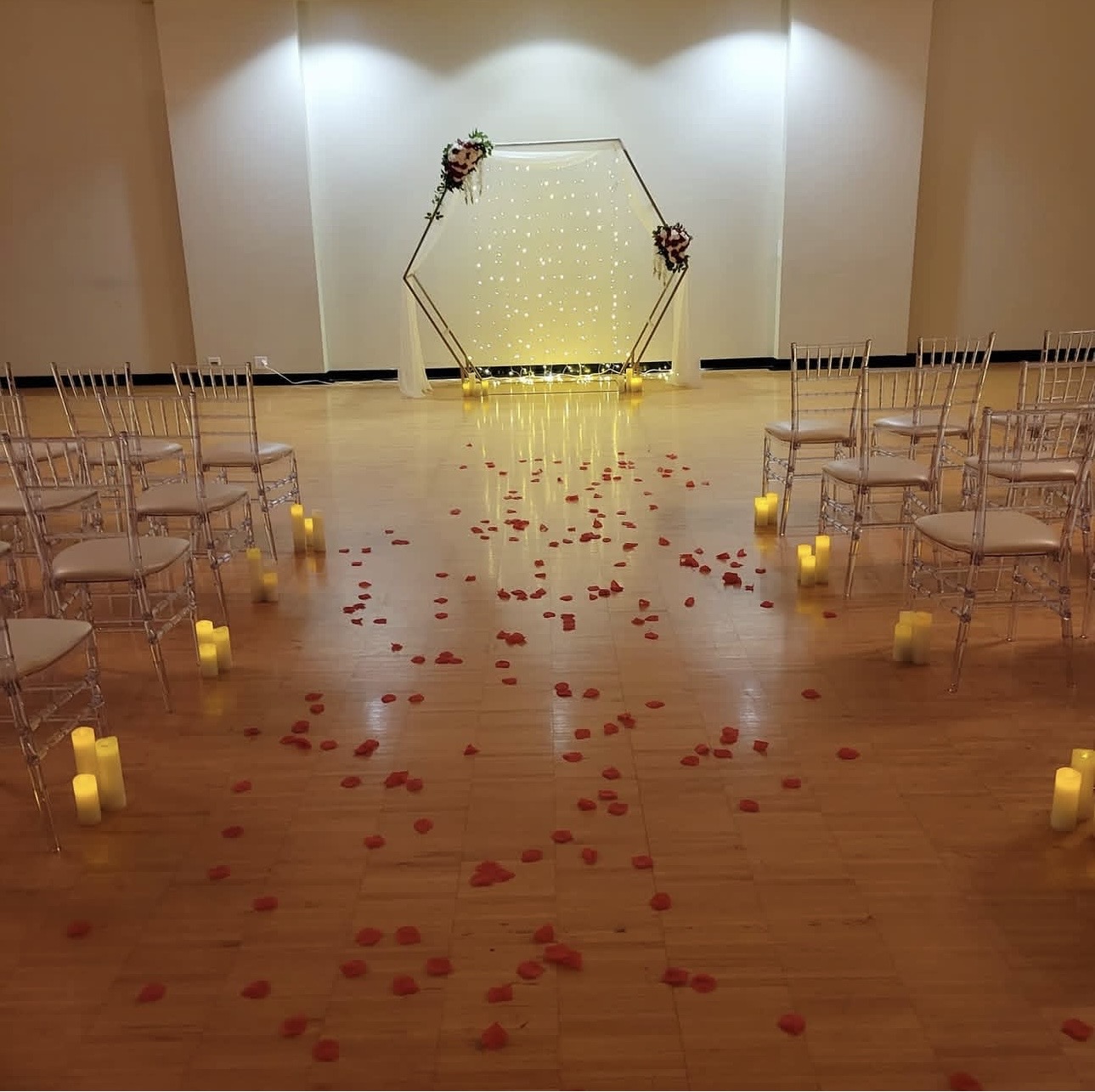 Wedding ceremony setup with chairs, rose petals, candles, and a decorated arch in a hall.