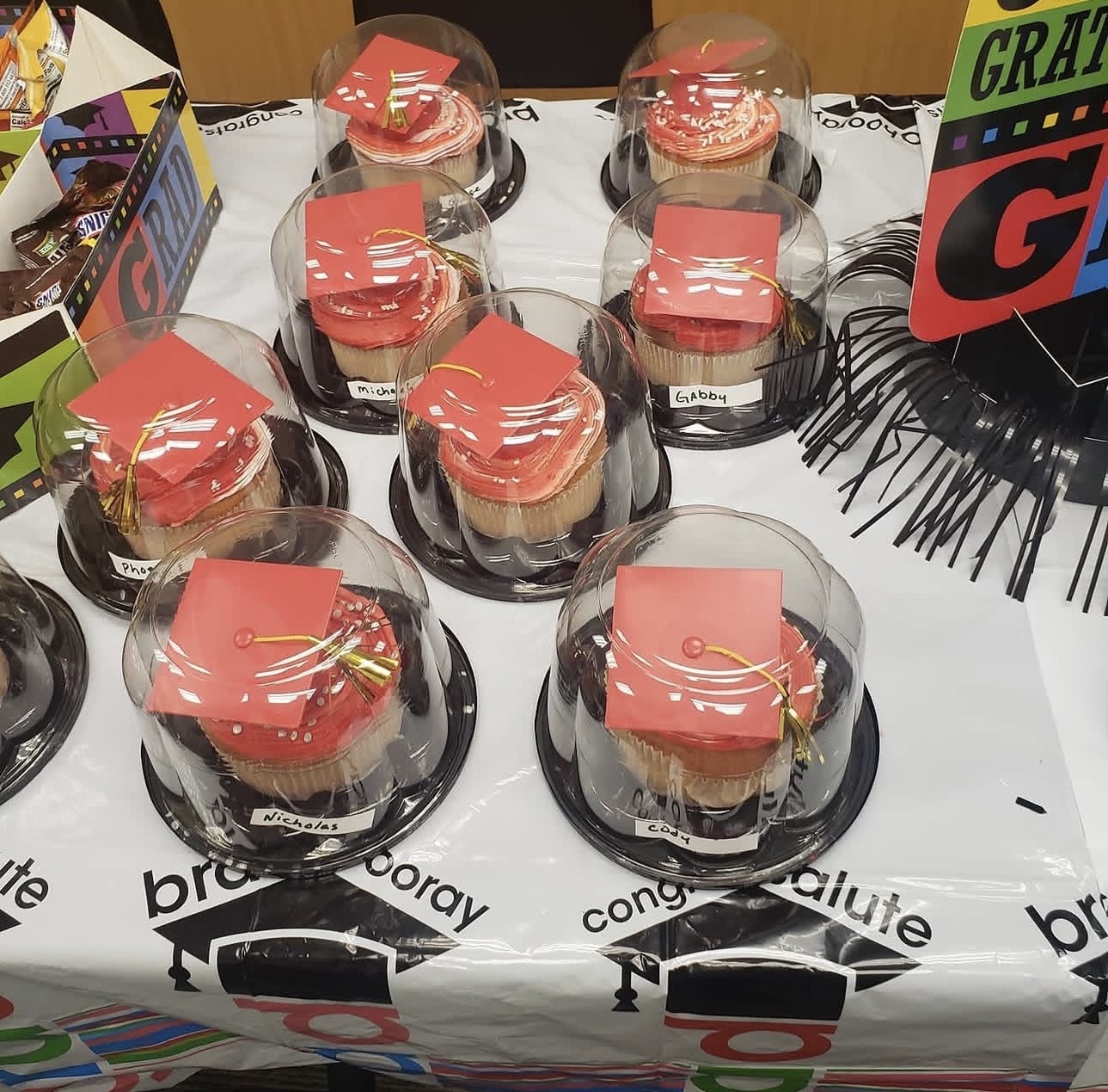 Six cupcakes with red icing and gold decorations inside clear plastic domes on a table, with colorful signs and decorations nearby.
