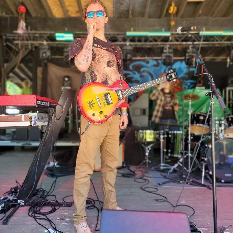 Person with sunglasses holding a yellow electric guitar on stage, with musical equipment and a drummer in the background.