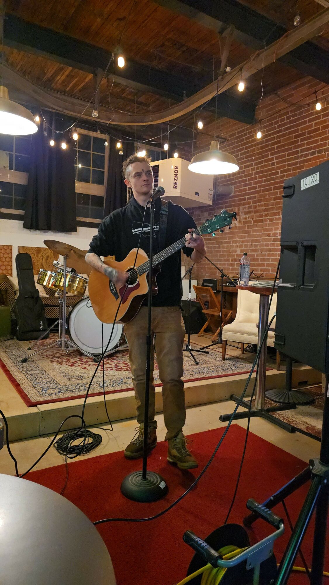 Person playing guitar in a room with musical equipment and a brick wall background.