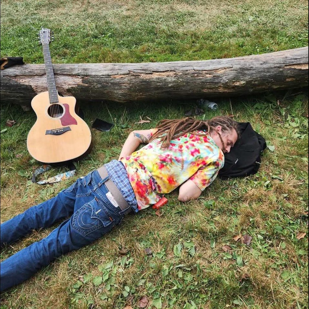 Person with dreadlocks lying on grass with a colorful shirt, resting head on a backpack, near a fallen log and an acoustic guitar.