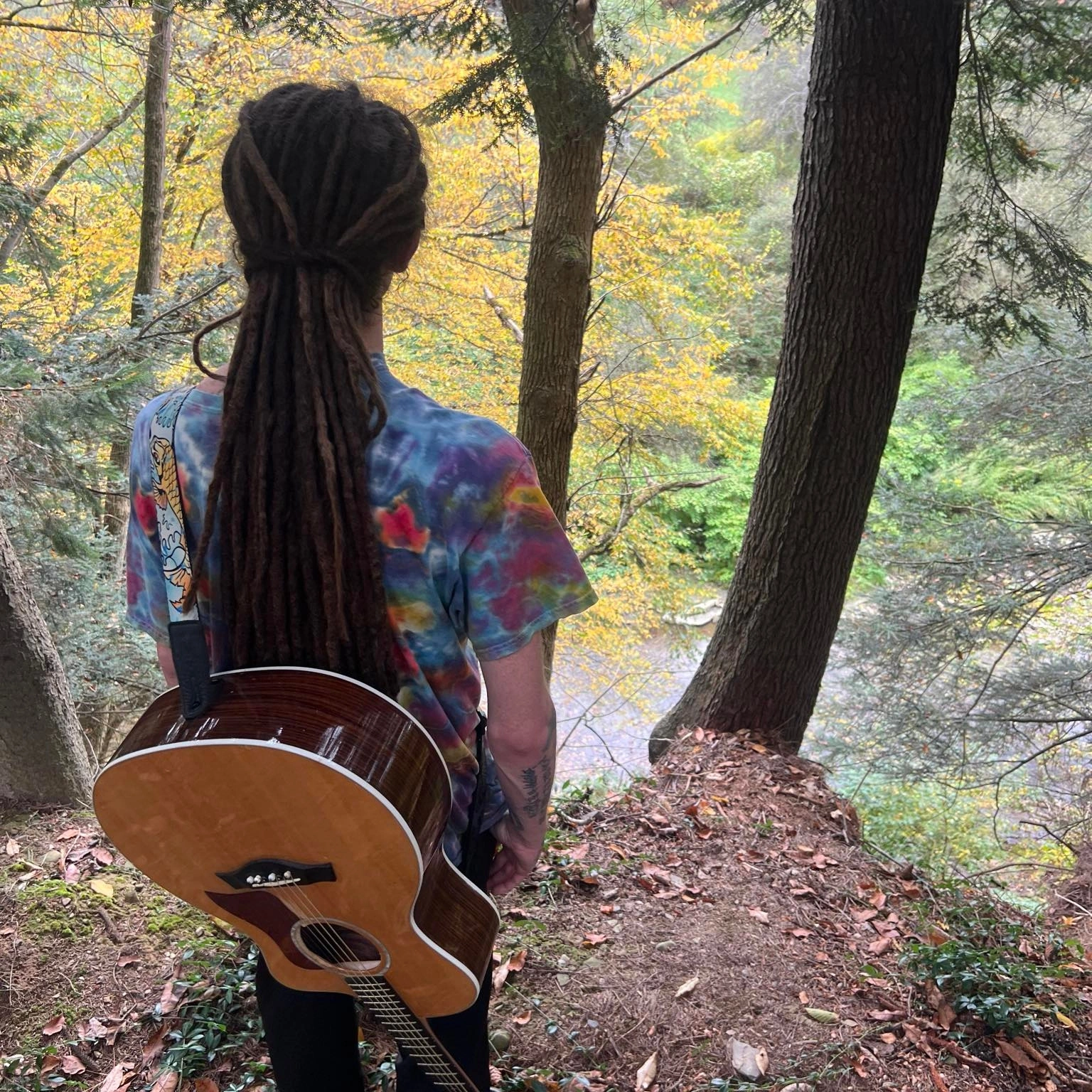 Person with long dreadlocks wearing a colorful shirt, standing in a forested area with trees and autumn leaves.