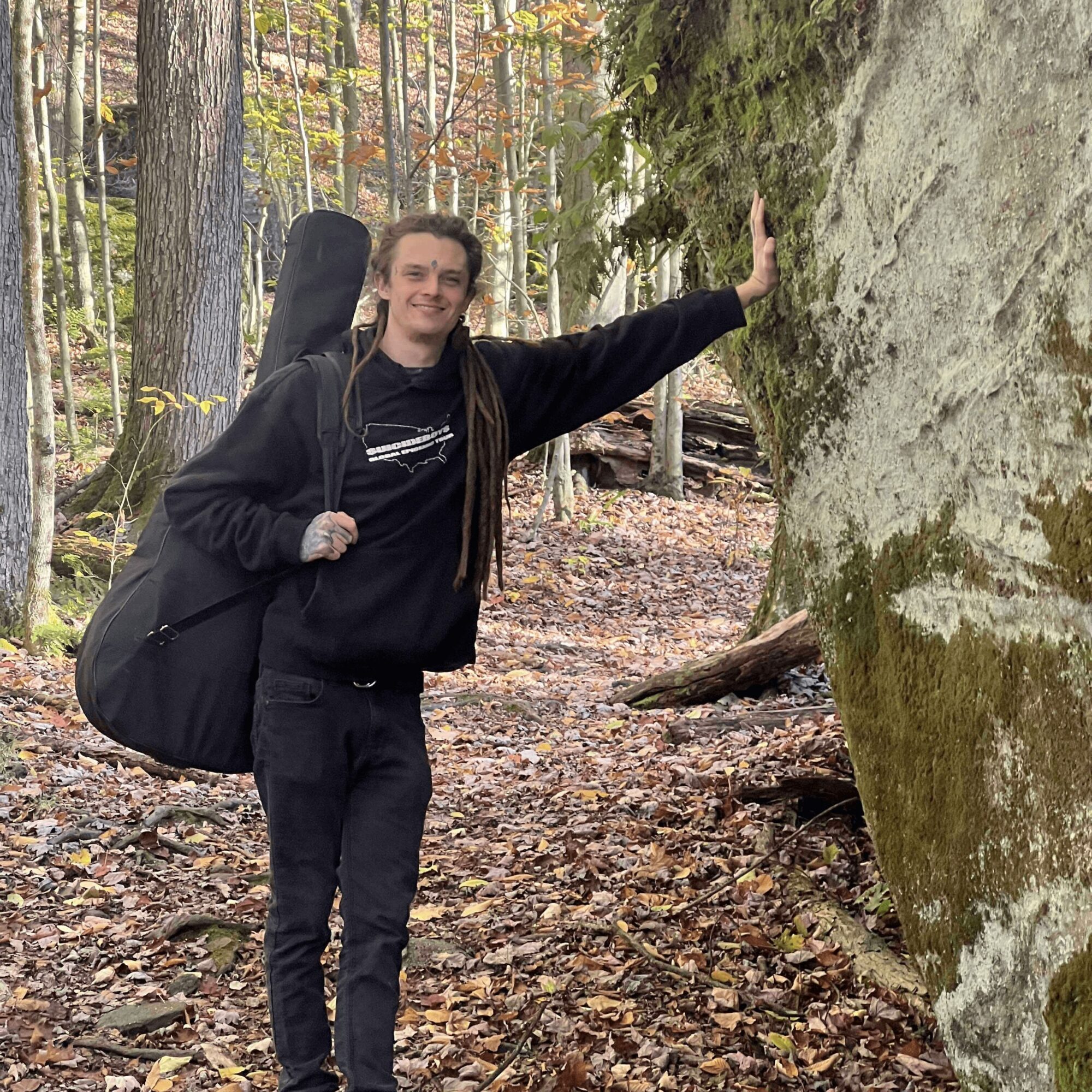 Person with long hair smiling, wearing a black hoodie, standing in a forest, touching a large mossy rock, carrying a guitar case.