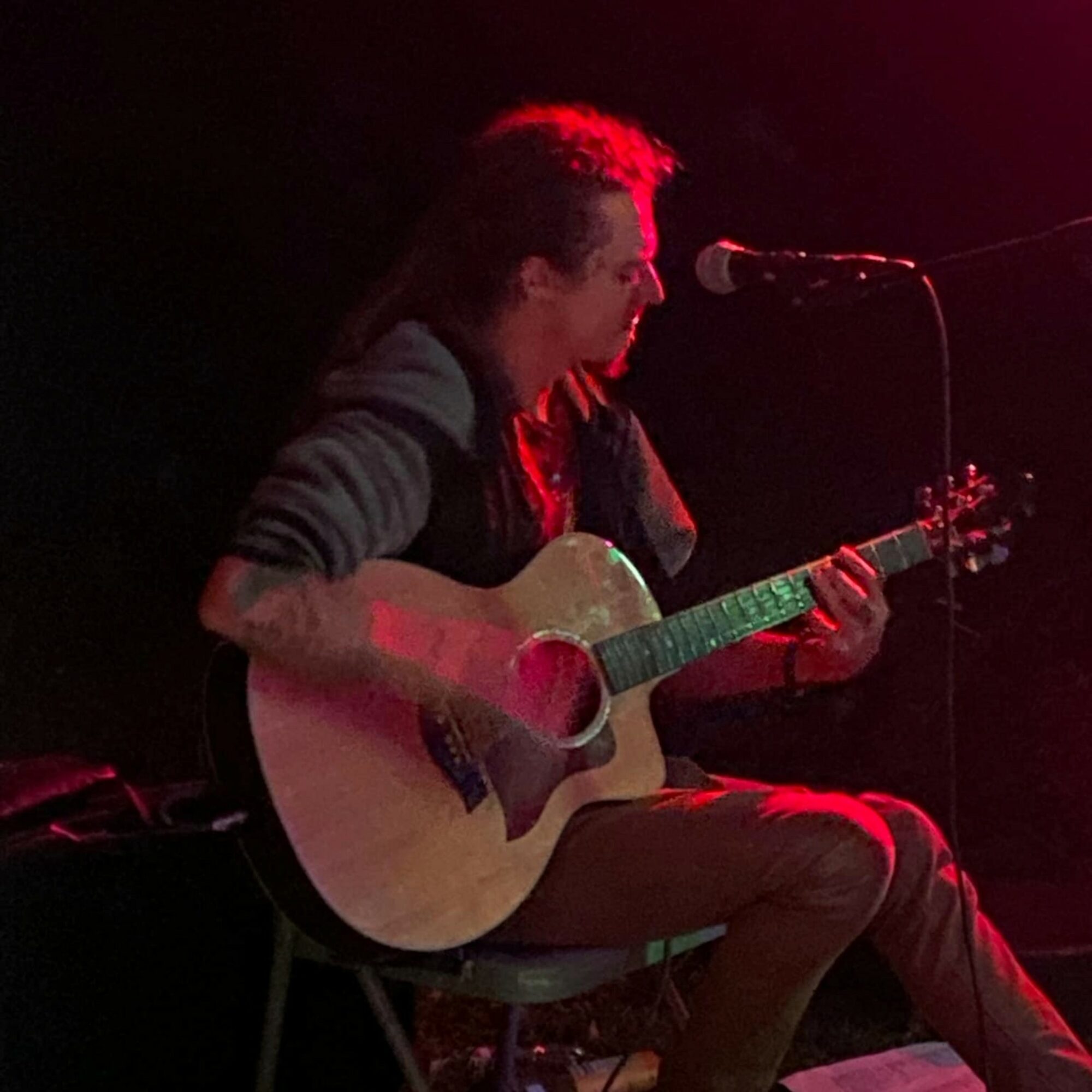 Person playing guitar and singing into microphone on stage, dim lighting with red hue, seated on chair.
